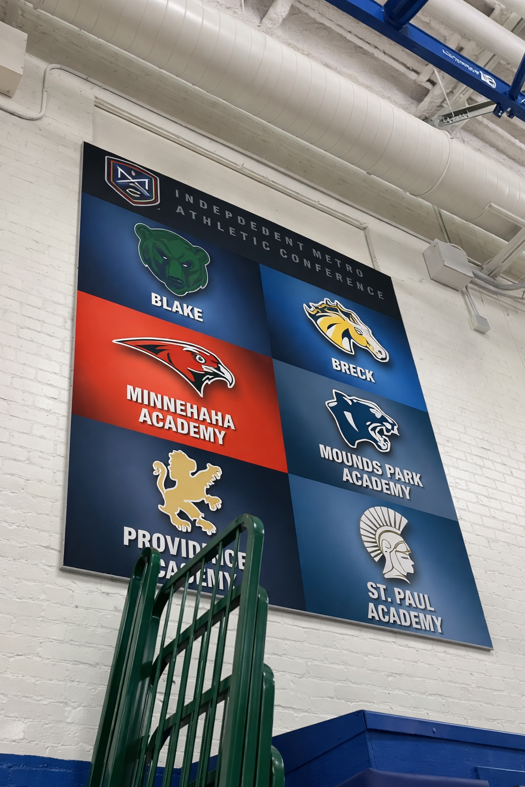 A large banner on a white brick wall displaying logos and names of teams competing in the Independent Metro Athletic Conference.