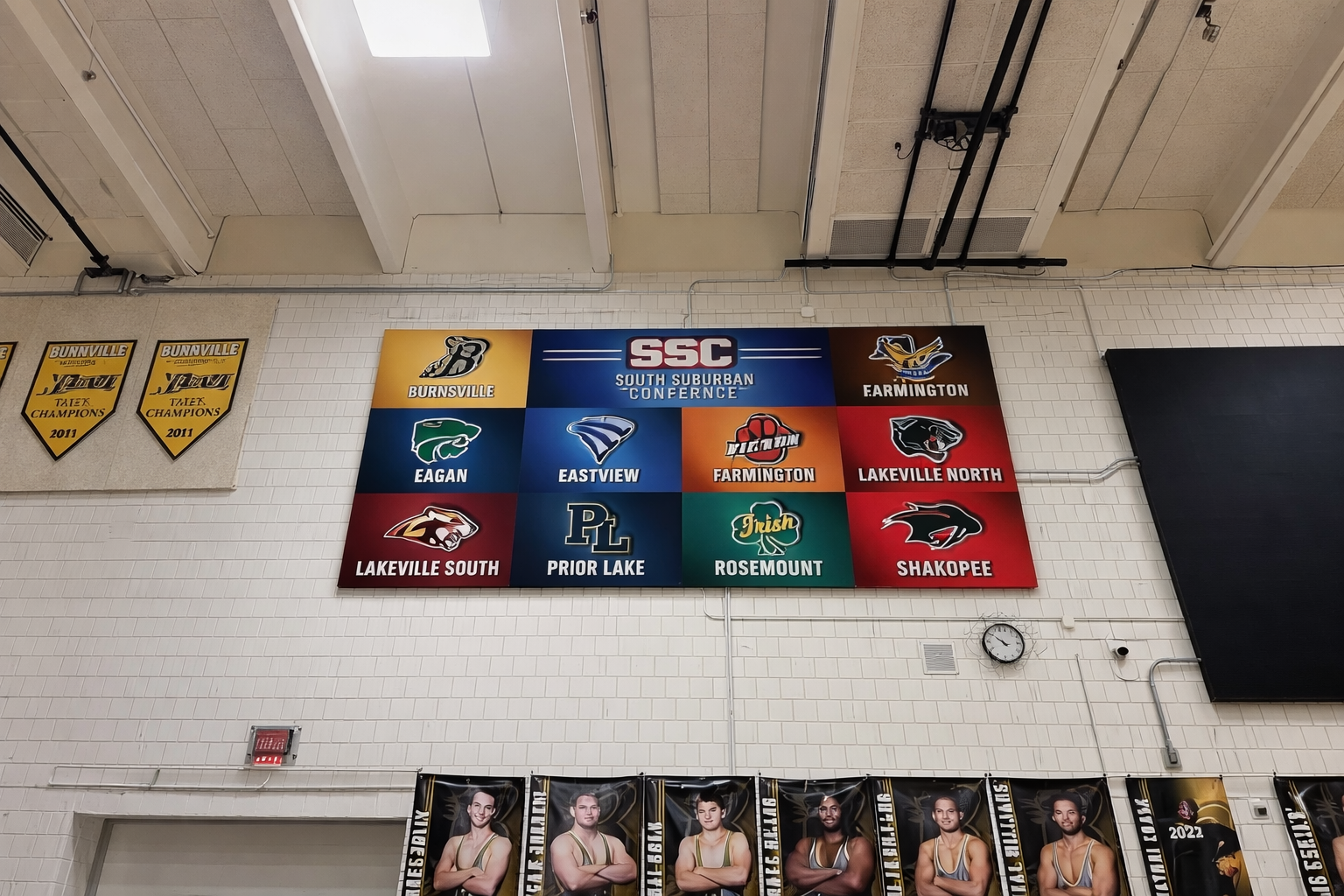 A large scoreboard displays the South Suburban Conference sports teams, with logos and names including Burnsville, Farmington, Eagan, Eastview, Lakeville North, Lakeville South, Prior Lake, Rosemount, and Shakopee. To the left, there are yellow banne