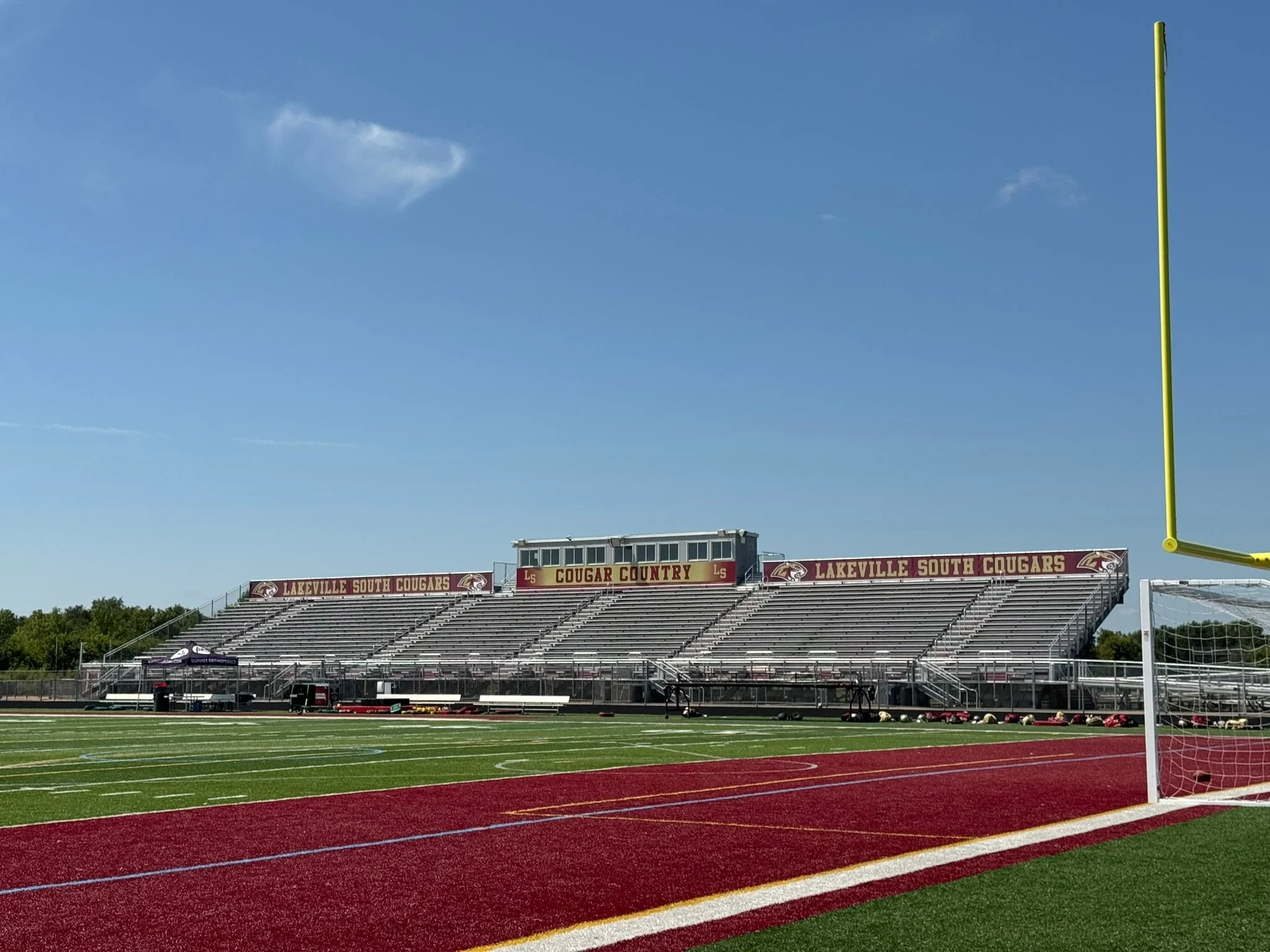 Photo of a football stadium with empty bleachers and a field, with a sign reading 'Lakeville South Cougars' and a football goal post on the right side.