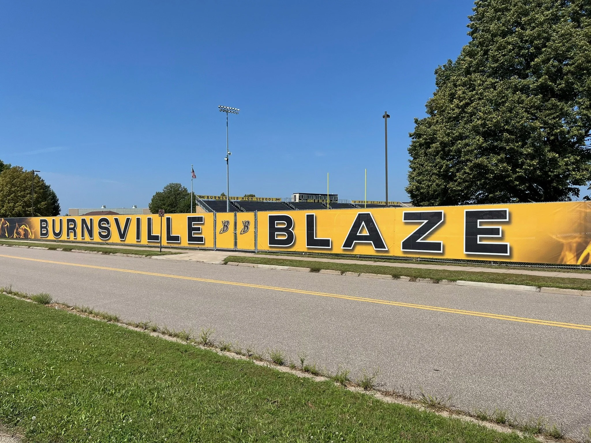 Yellow banner reading "Burnsville Blaze" with the school's football field and bleachers in the background, sunny day with blue sky and trees.