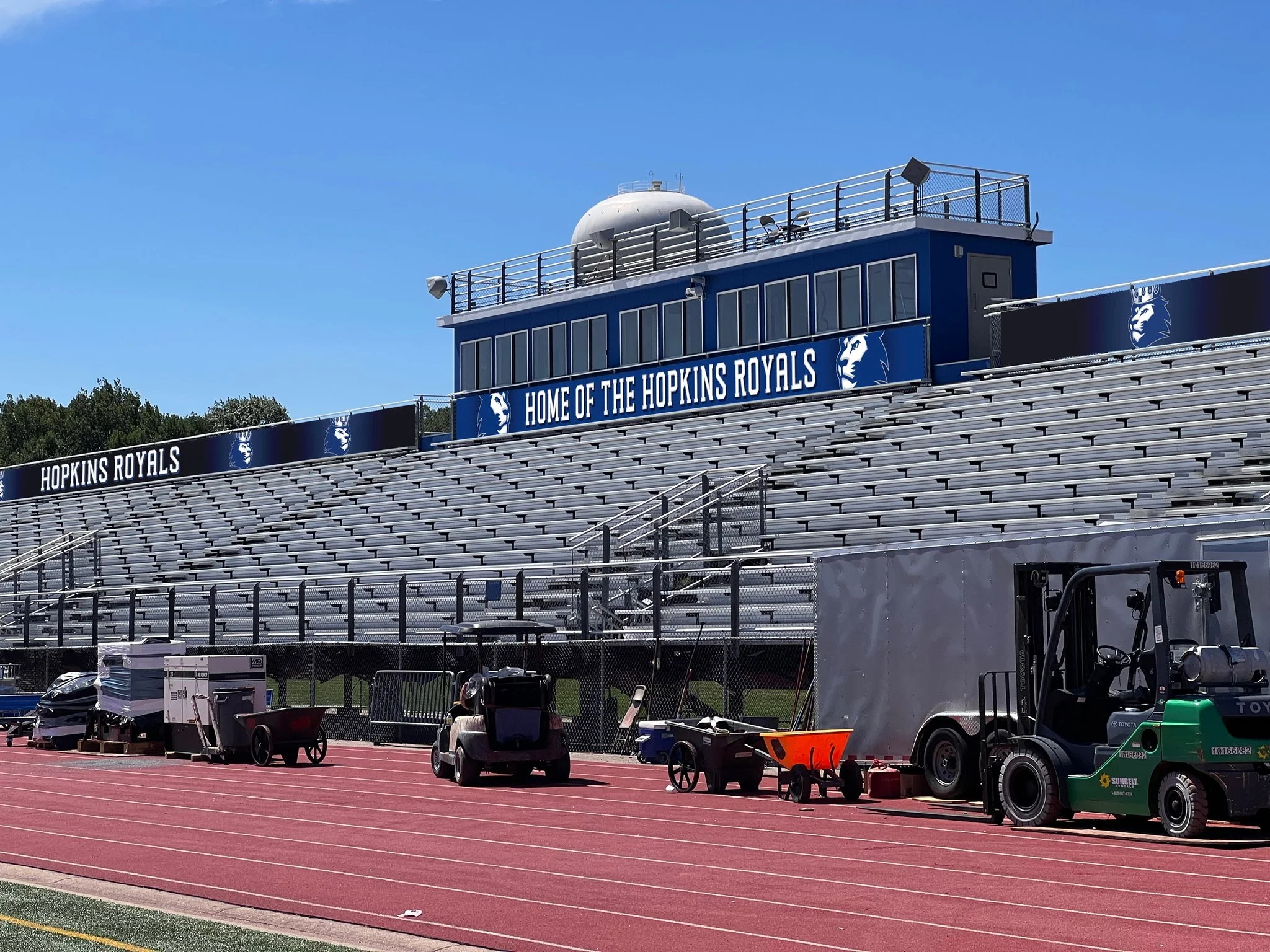 Empty football stadium bleachers with signage reading 'Home of the Hopkins Royals'. The field side has equipment and carts, and clear skies overhead.