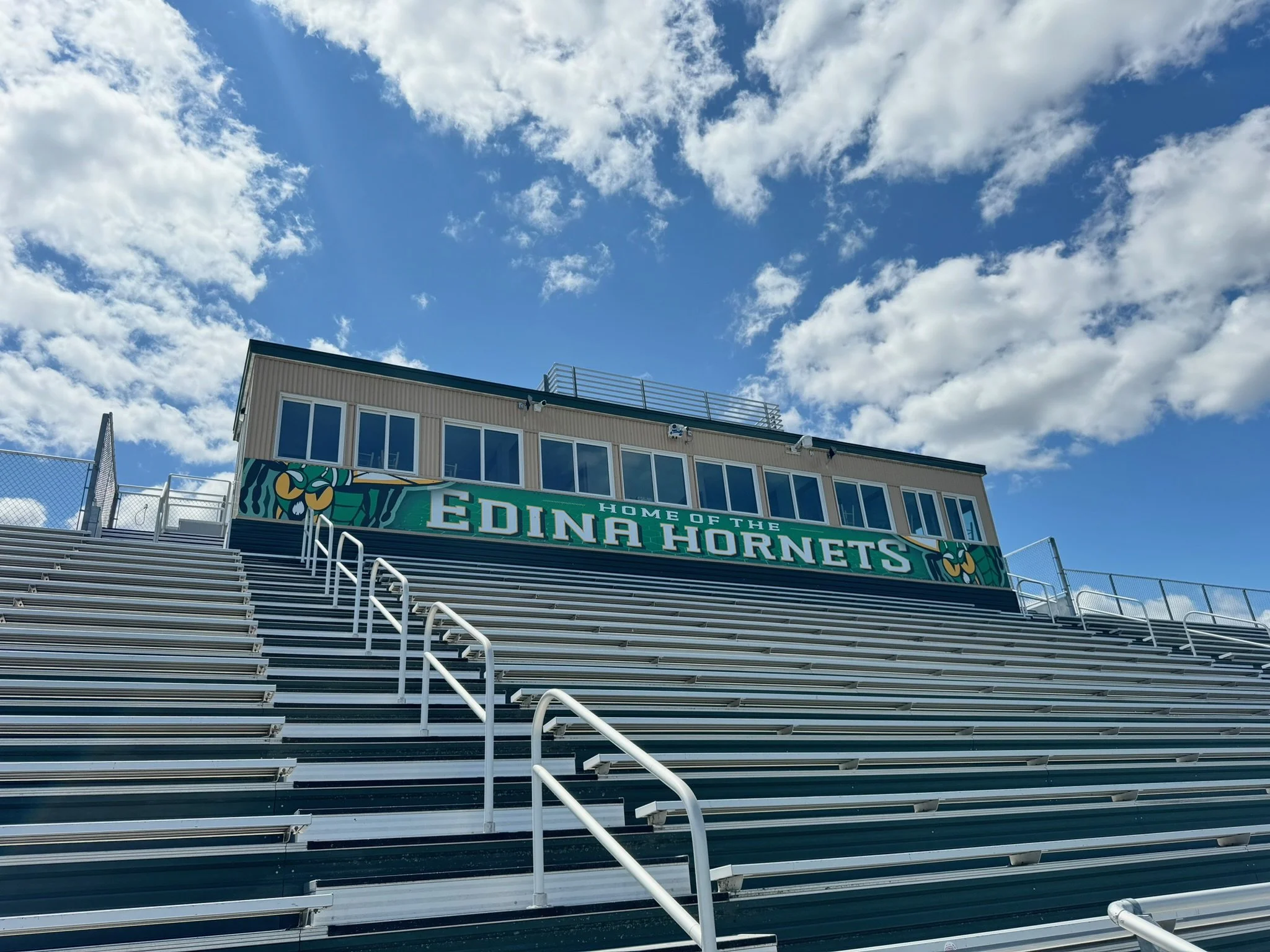 Empty stadium bleachers with a sign that reads 'Home of the Edina Hornets' under a partly cloudy sky.