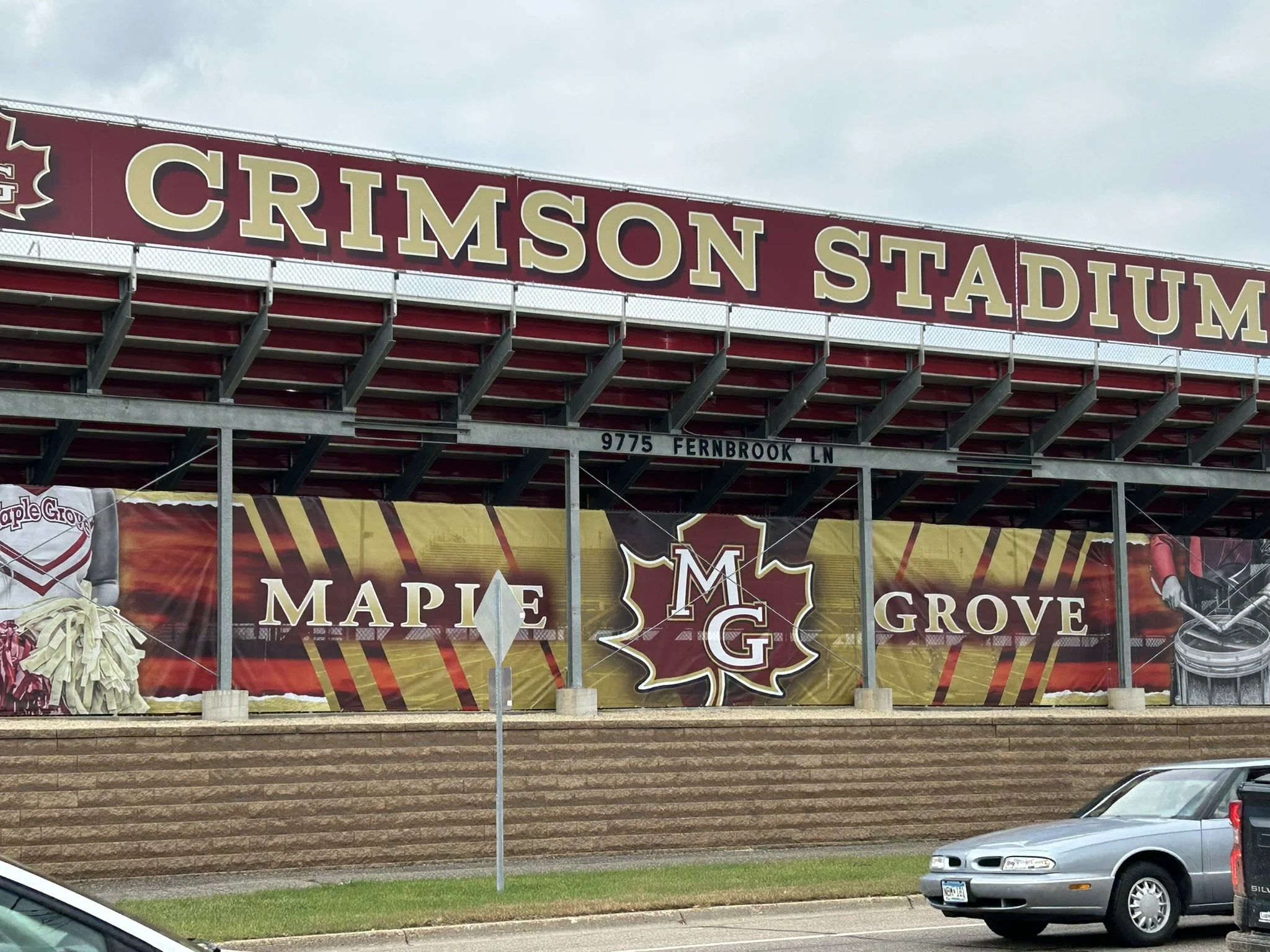 Exterior view of Crimson Stadium showing a large sign with the words 'Maple Grove' and the initials 'MG' on a maple leaf emblem, along with a football helmet and pom-poms.