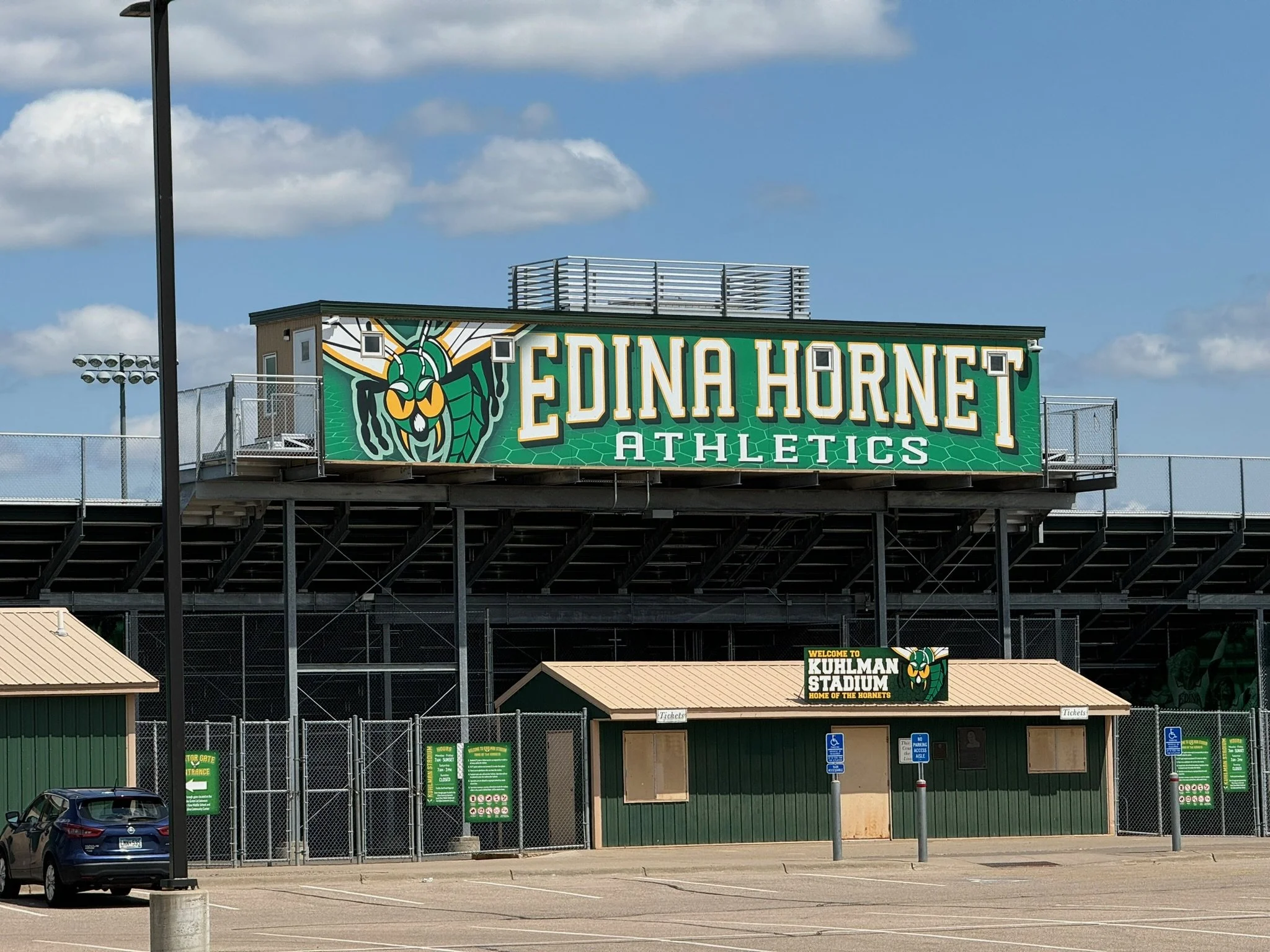 Exterior view of Edina Hornet Athletics at Kuhlman Stadium, showing a large green sign with a hornet mascot, parking area, and stadium entrance with a smaller sign and handicap parking signs.
