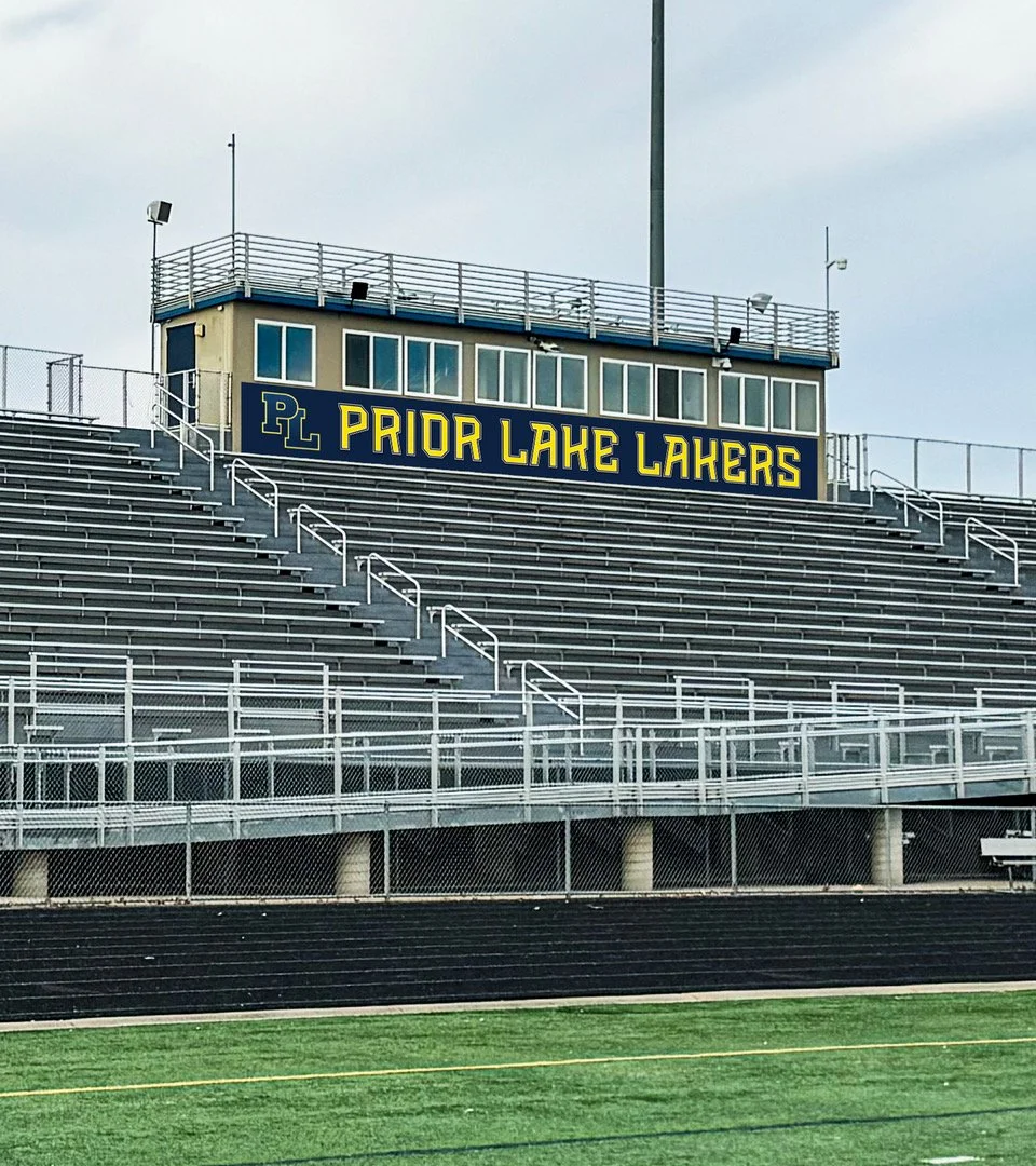 Empty bleachers at a sports stadium with a sign that reads 'PL PRIOR LAKE LAKERS' above a small press box.