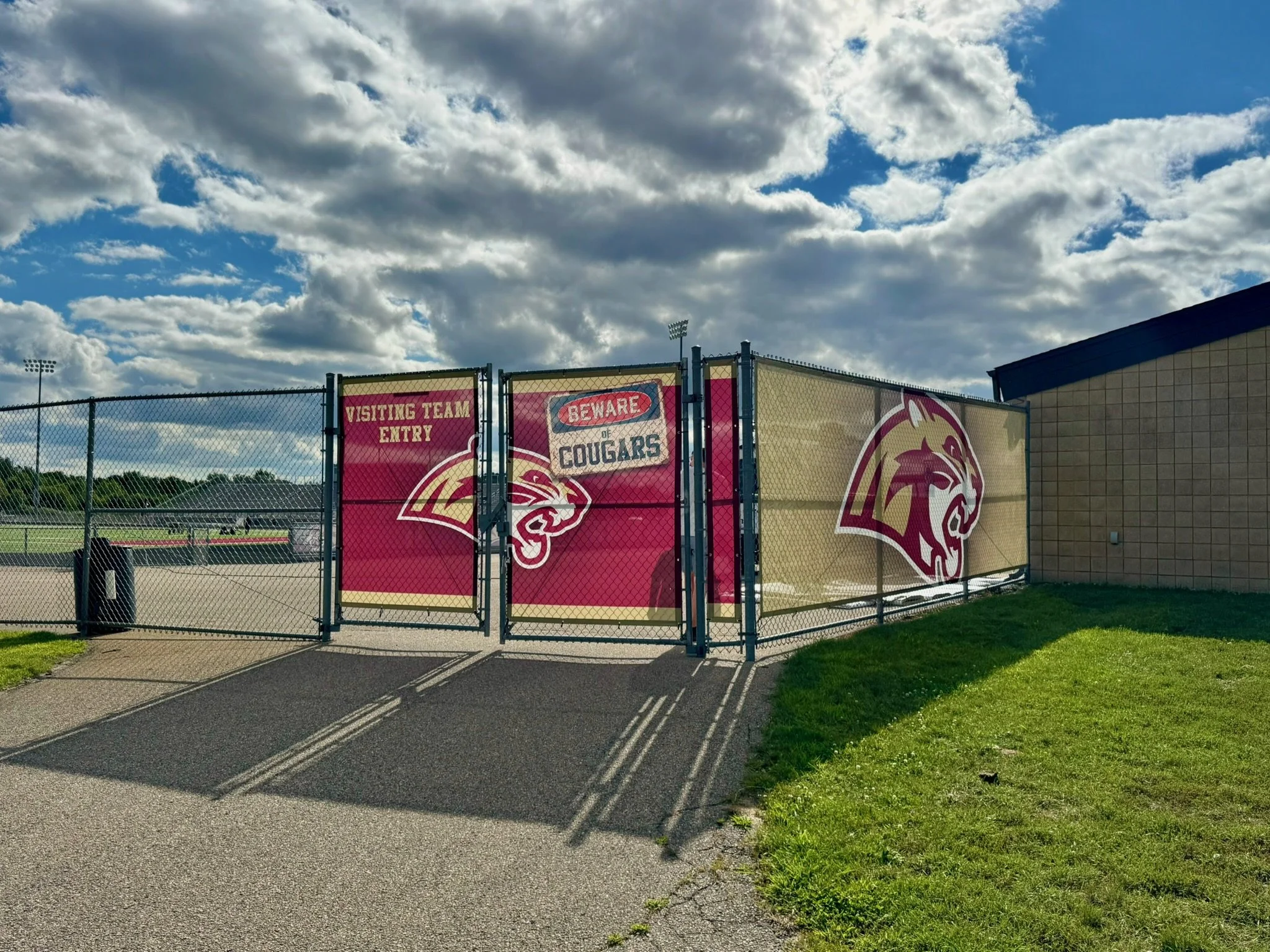 Football field entrance gate with signs reading 'Visiting Team Entry' and 'Beware Cougars,' featuring a cougar mascot logo, under a partly cloudy sky.