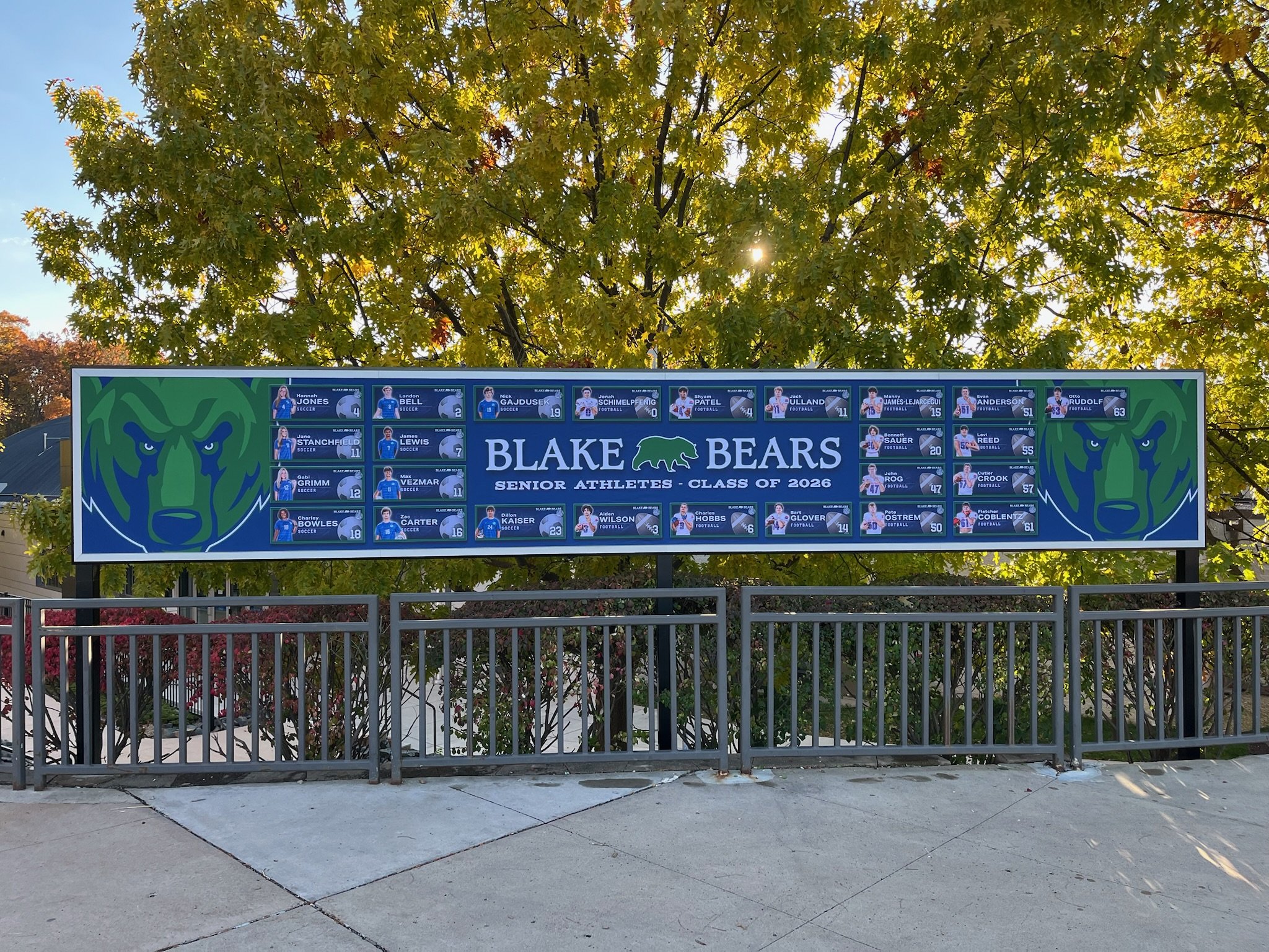 School sign honoring Blake Bears senior athletes from the class of 2026, featuring student portraits, names, and numbers, with a large blue bear head logo on either side, set under a tree with green leaves.