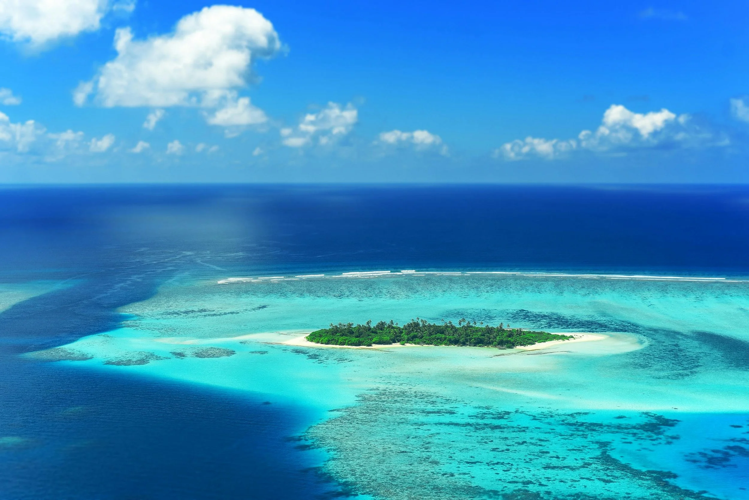 A tropical island surrounded by turquoise waters and coral reefs, with a dense green forest, under a partly cloudy blue sky.
