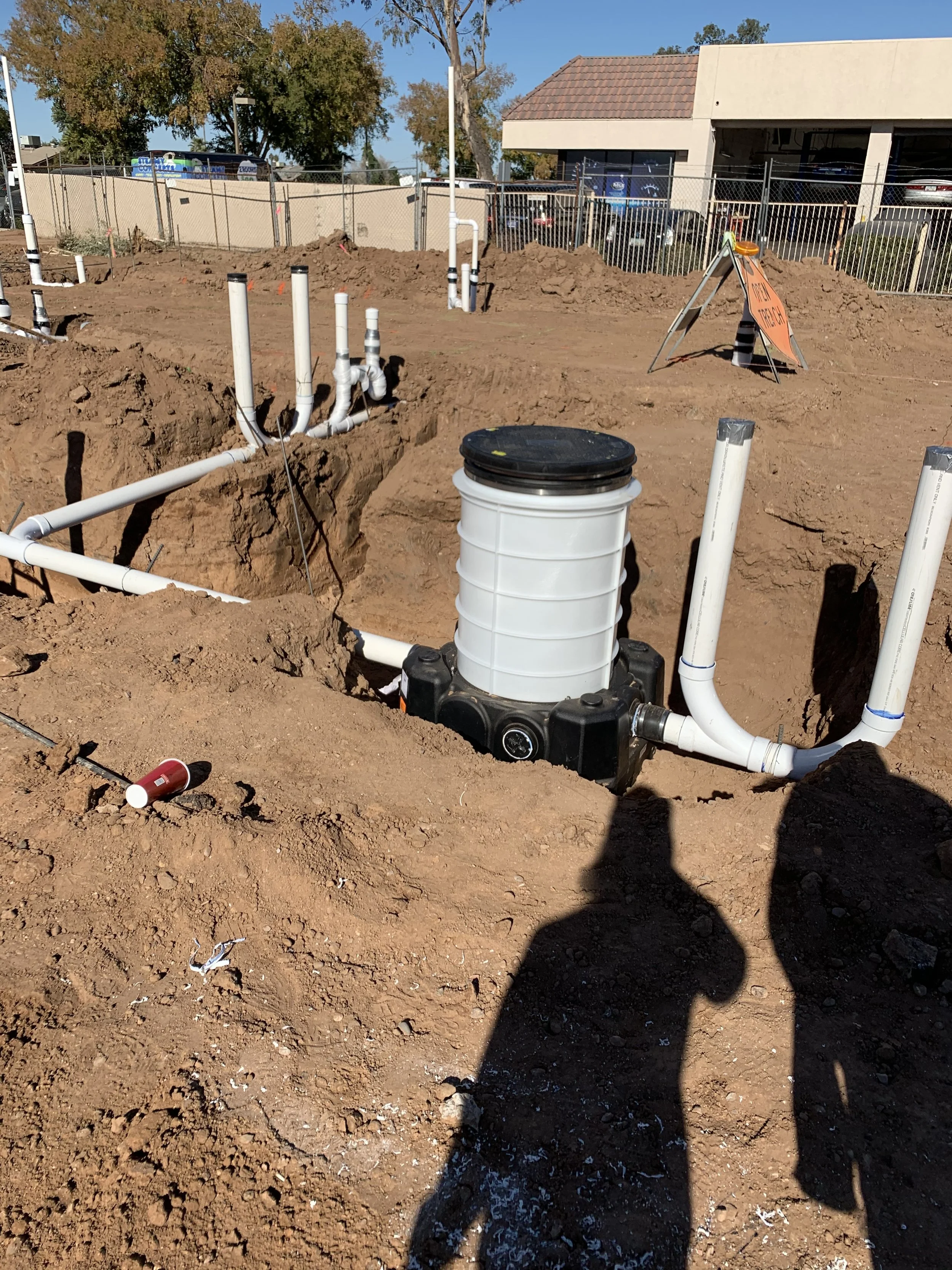 Construction site with exposed plumbing pipes and a large white filter or pump in the center, surrounded by dirt and construction markers, with a building and trees in the background.