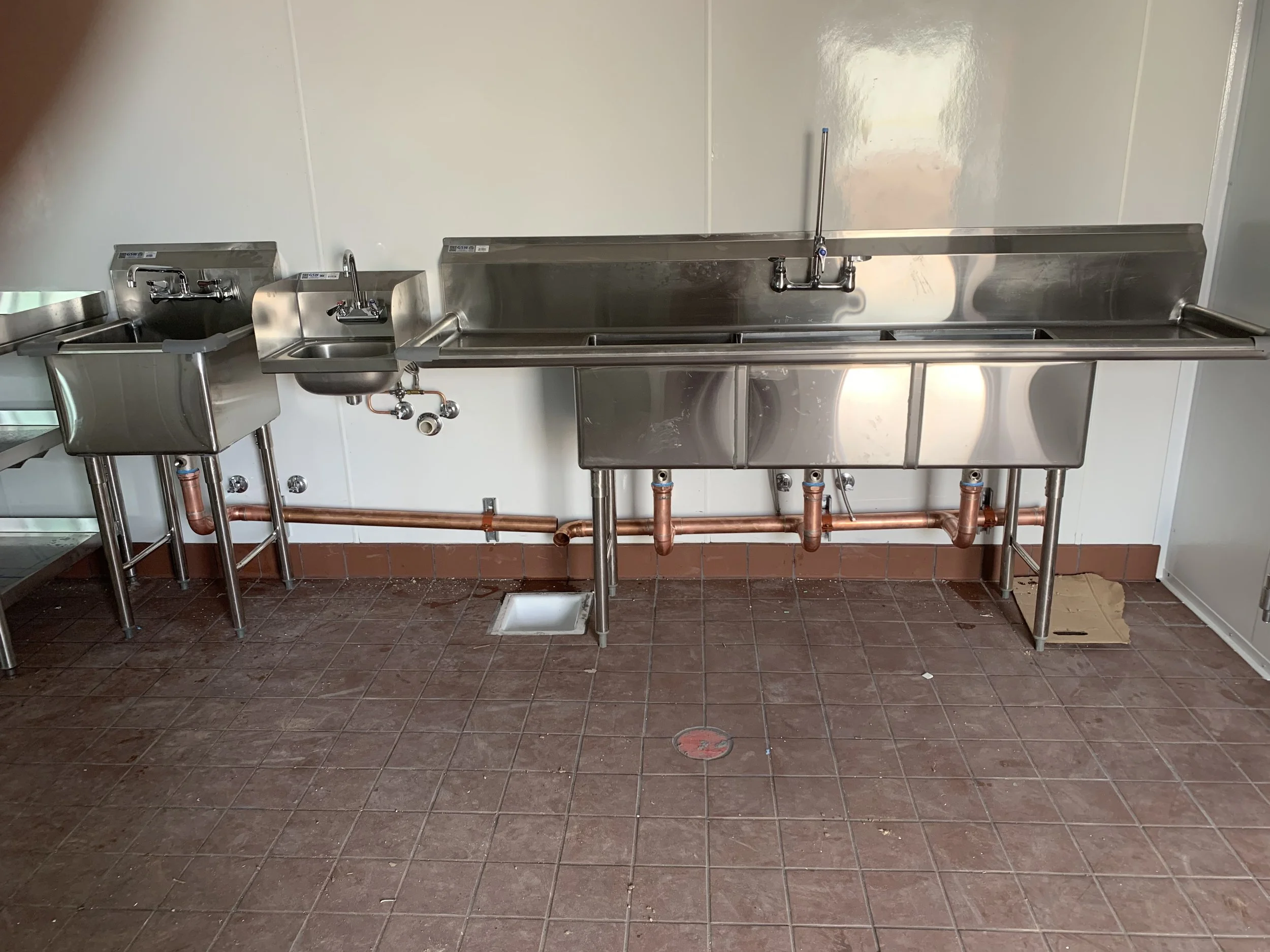 A clean metal utility sink with a hand spray faucet, above a copper pipe drainage system, set against a white wall in a room with brown tile flooring.