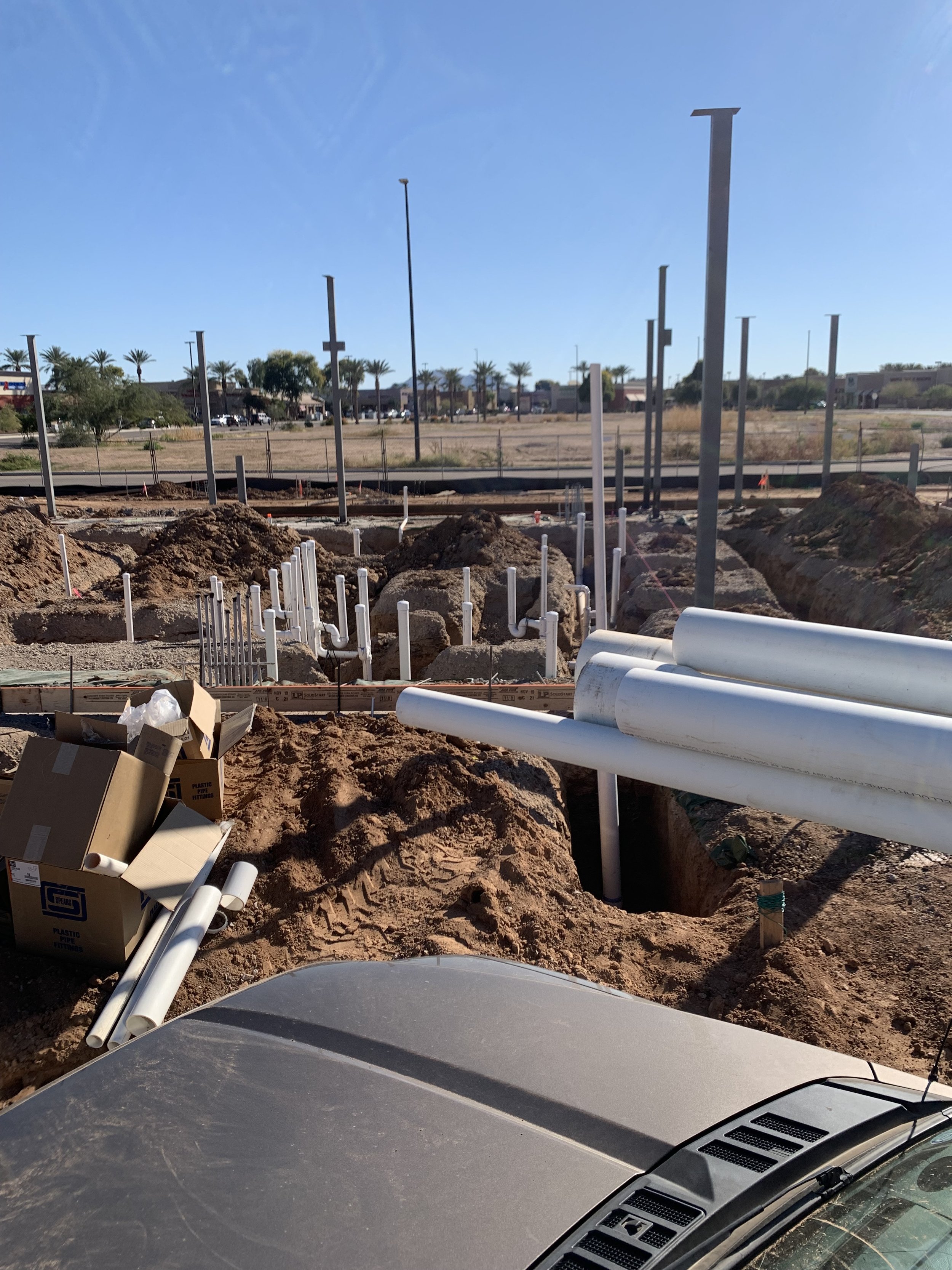 Construction site with dirt, white pipes, and metal poles in the ground, with a parking lot and palm trees in the background under a blue sky.