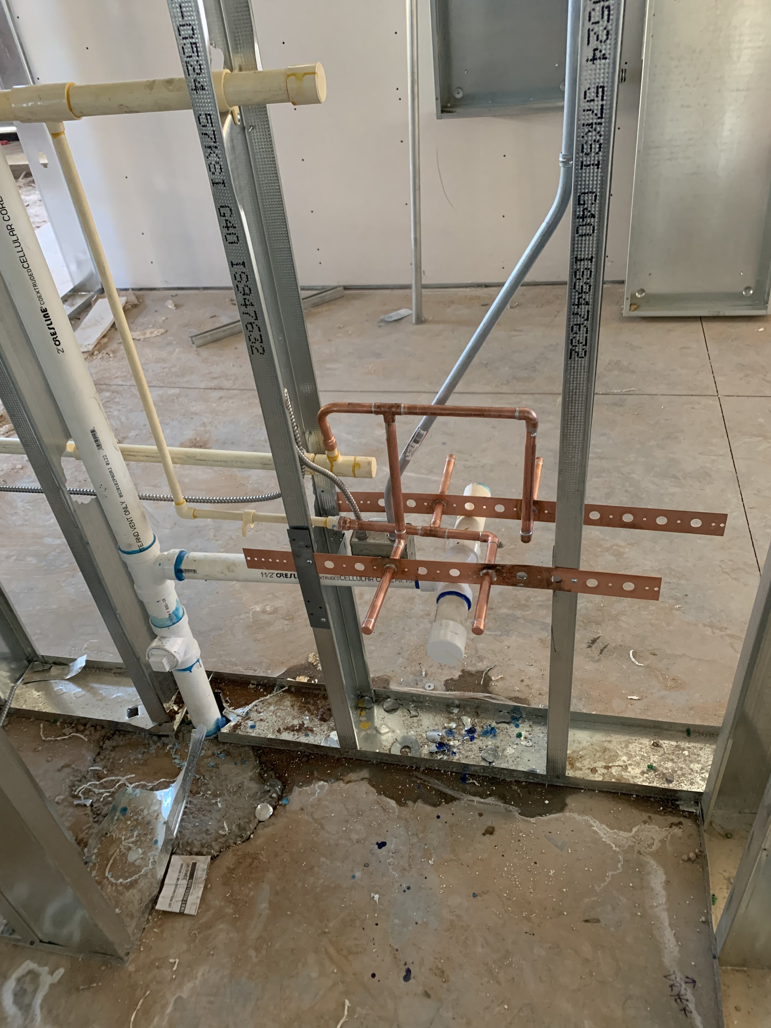 Interior view of a construction site with exposed plumbing and electrical wiring. Visible copper pipes, white plastic pipes, metal studs, and some debris on the floor.