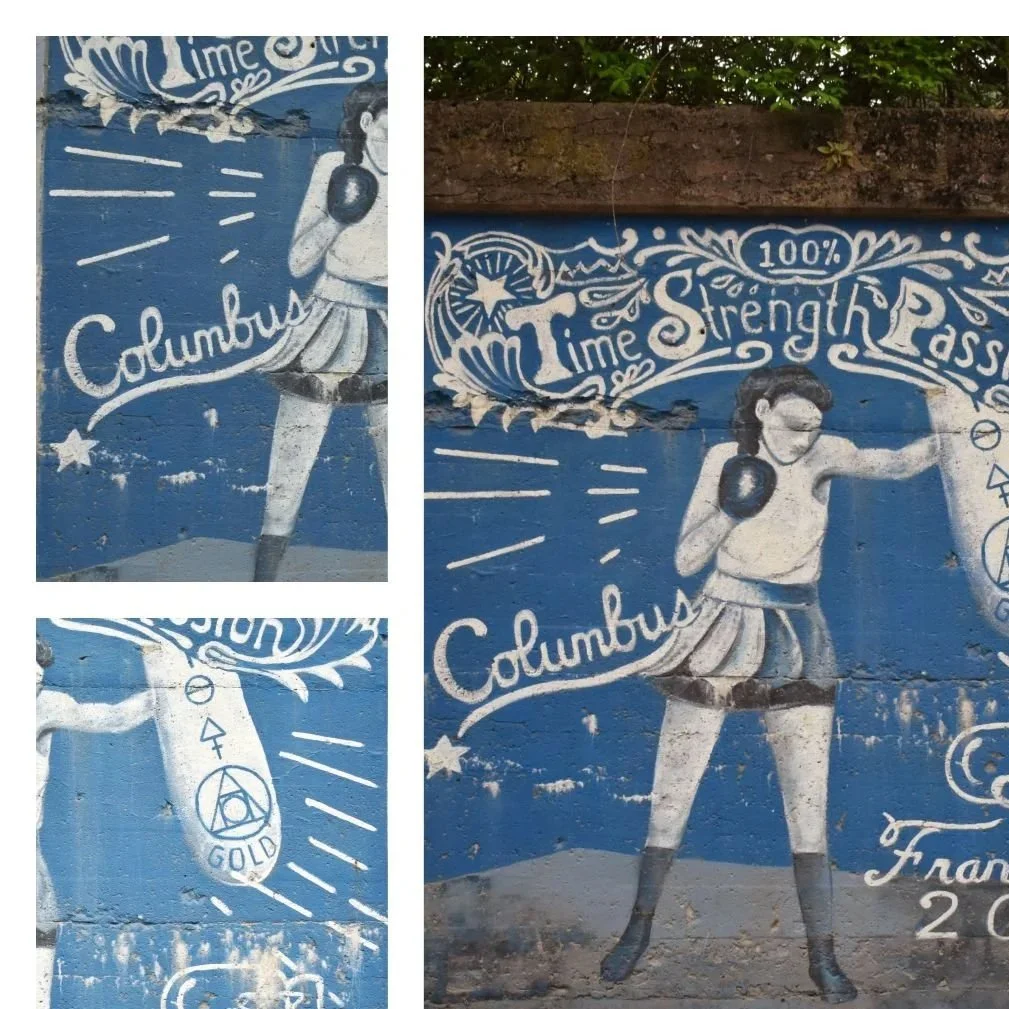 Street art mural of a female boxer in a boxing stance, wearing boxing gloves and shorts, with decorative text and symbols surrounding her, including phrases like 'Time,' 'Strength,' and 'Class.'