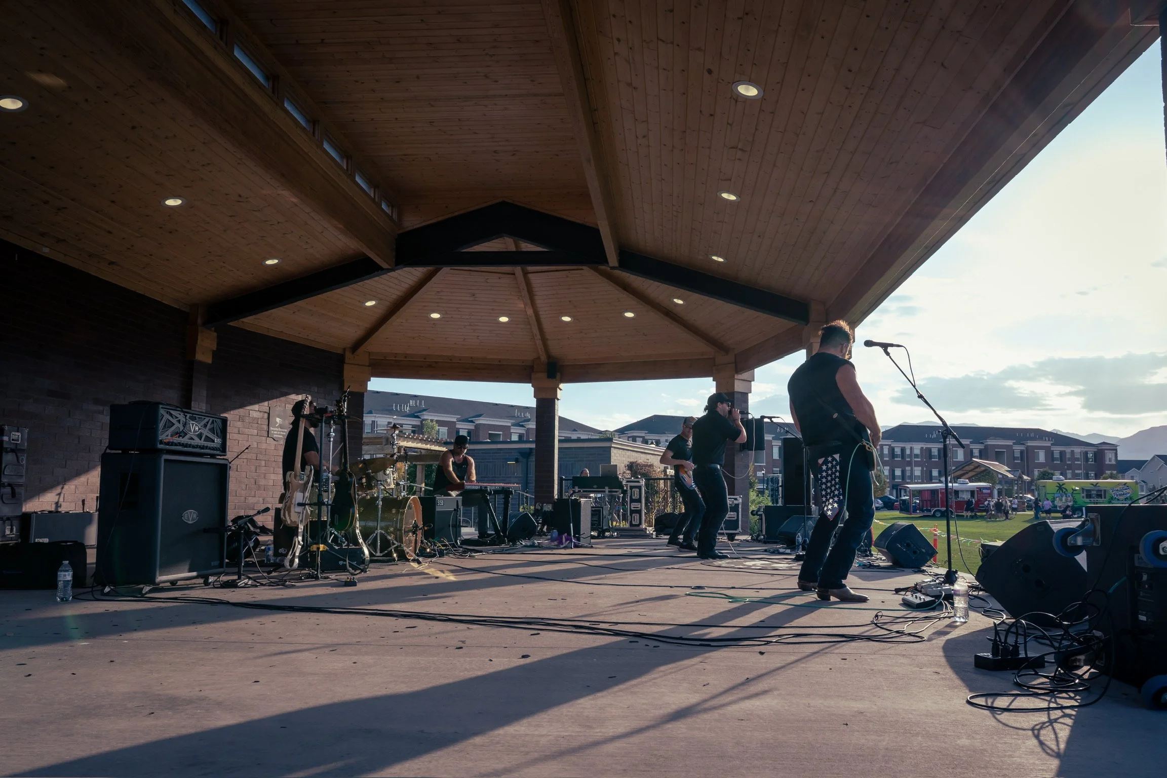 A band performing on an outdoor stage with wooden roof, during sunset in a neighborhood park.