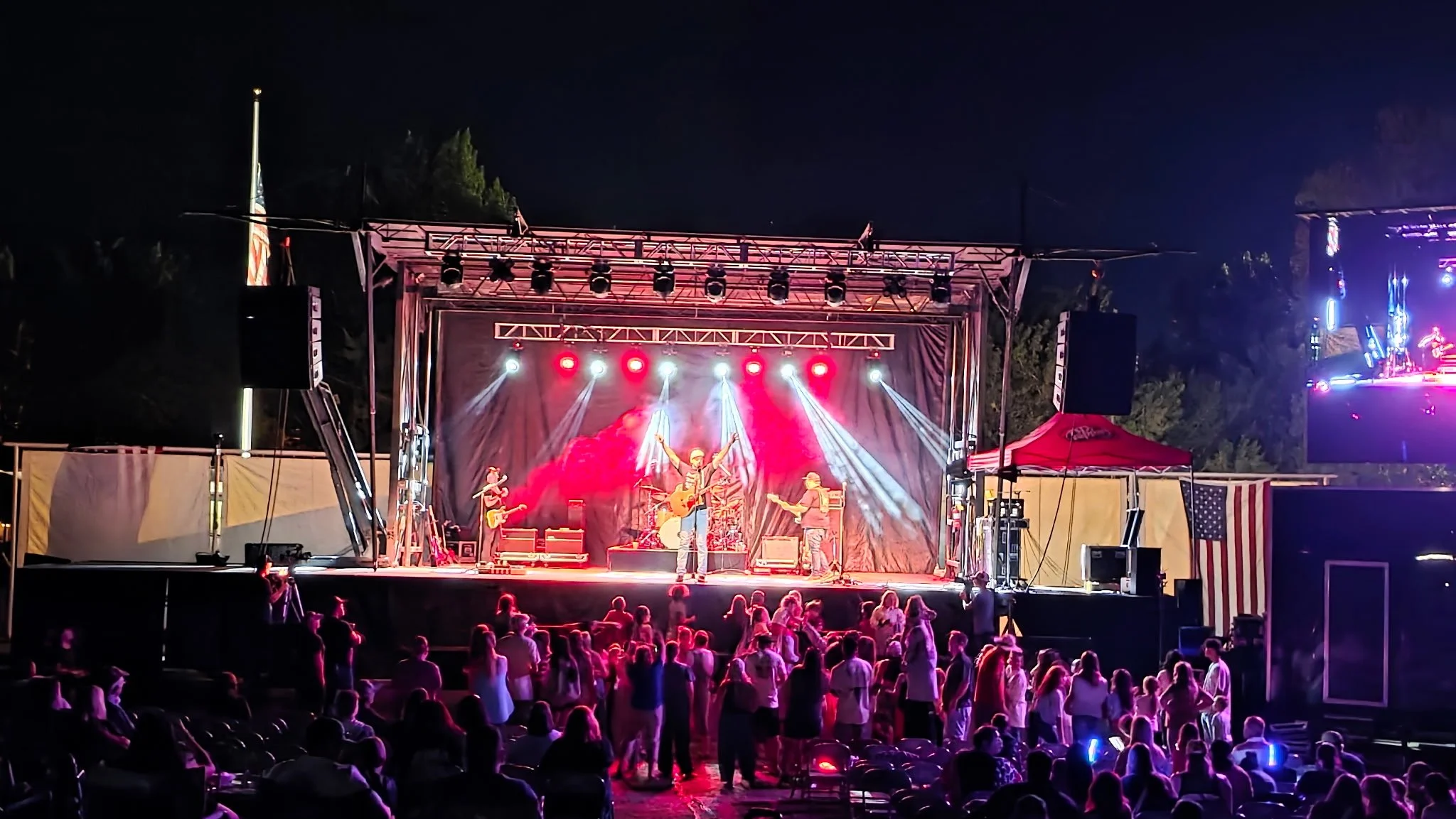 Live outdoor music concert at night with a band performing on stage, audience standing and sitting, stage lights shining, American flag on the right side.