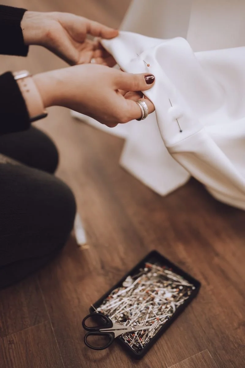 Person sewing a white fabric, possibly a garment, with a tray of pins and small scissors nearby on a wooden floor.