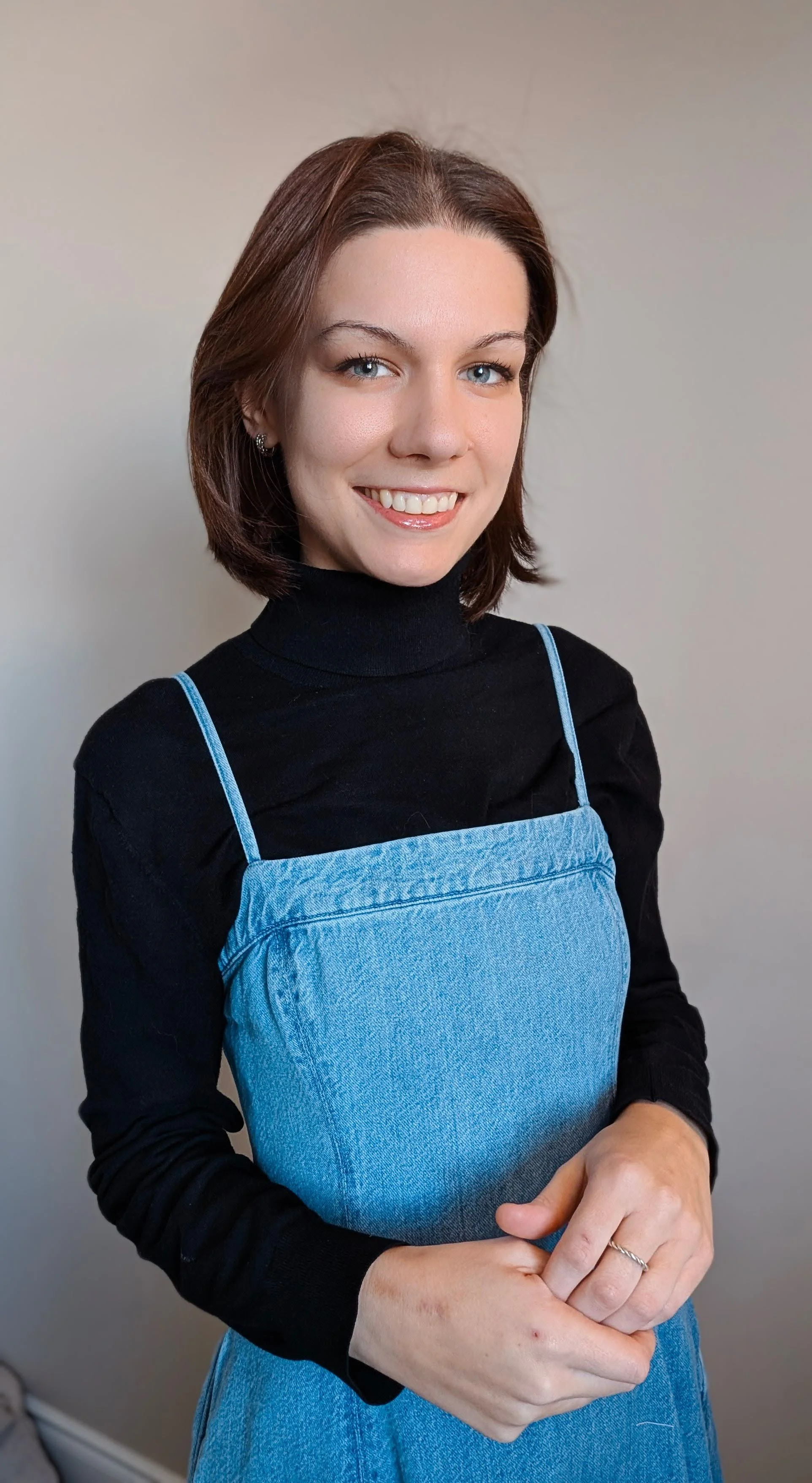 A young woman with shoulder-length brown hair, blue eyes, and light skin, smiling and looking at the camera, wearing a black turtleneck and a blue denim overall dress.