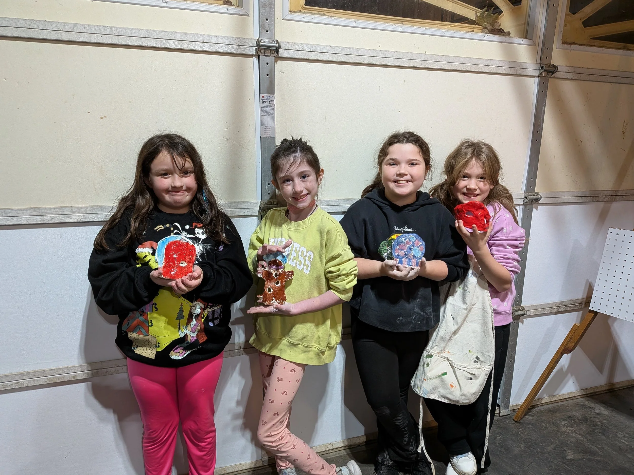 Four young girls standing side by side indoors, each holding colorful handcrafted ceramic sculptures, smiling at the camera.