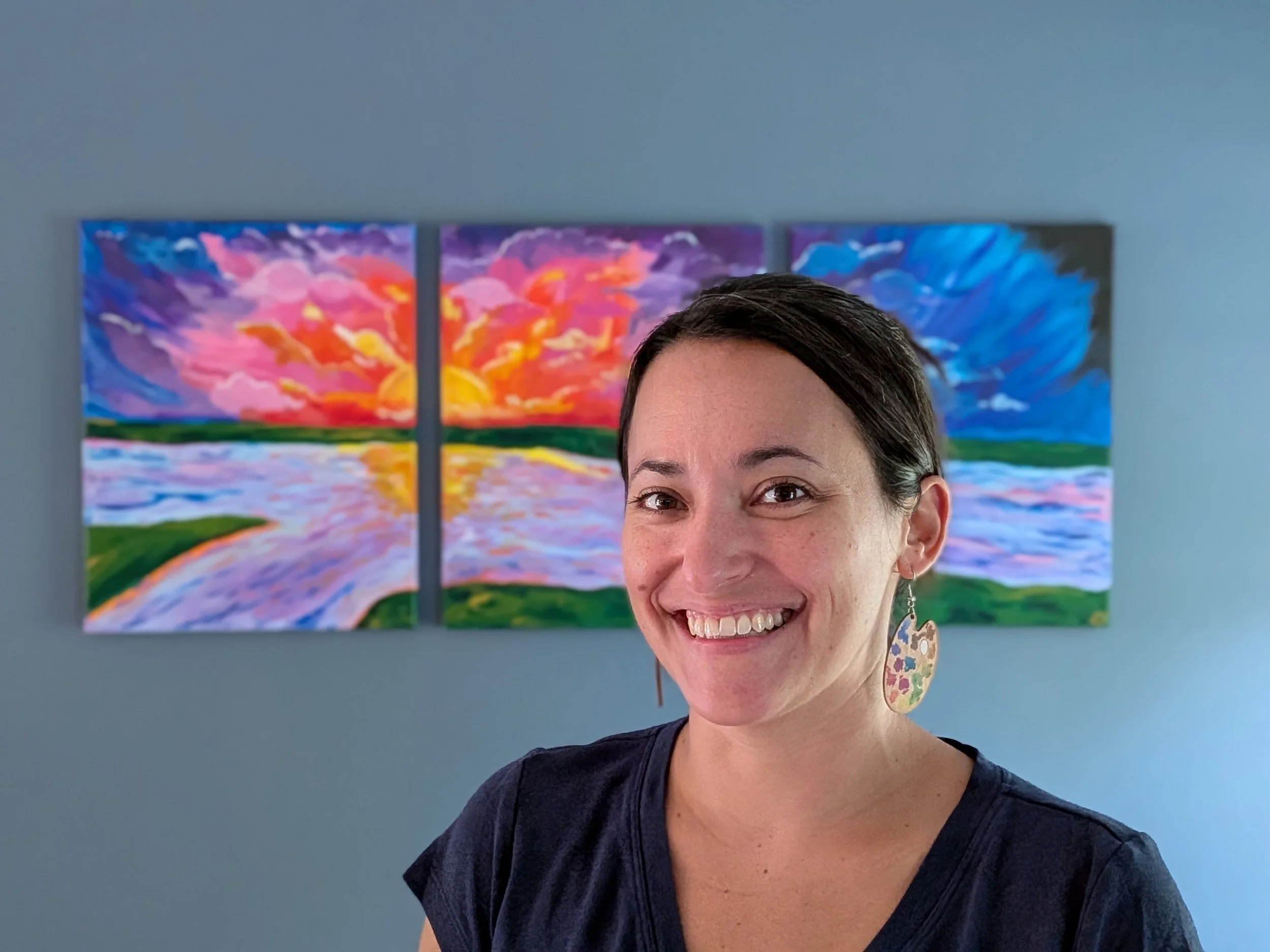 A woman smiling in front of a colorful triptych landscape painting depicting a sunset over water with purple, pink, orange, and blue clouds.