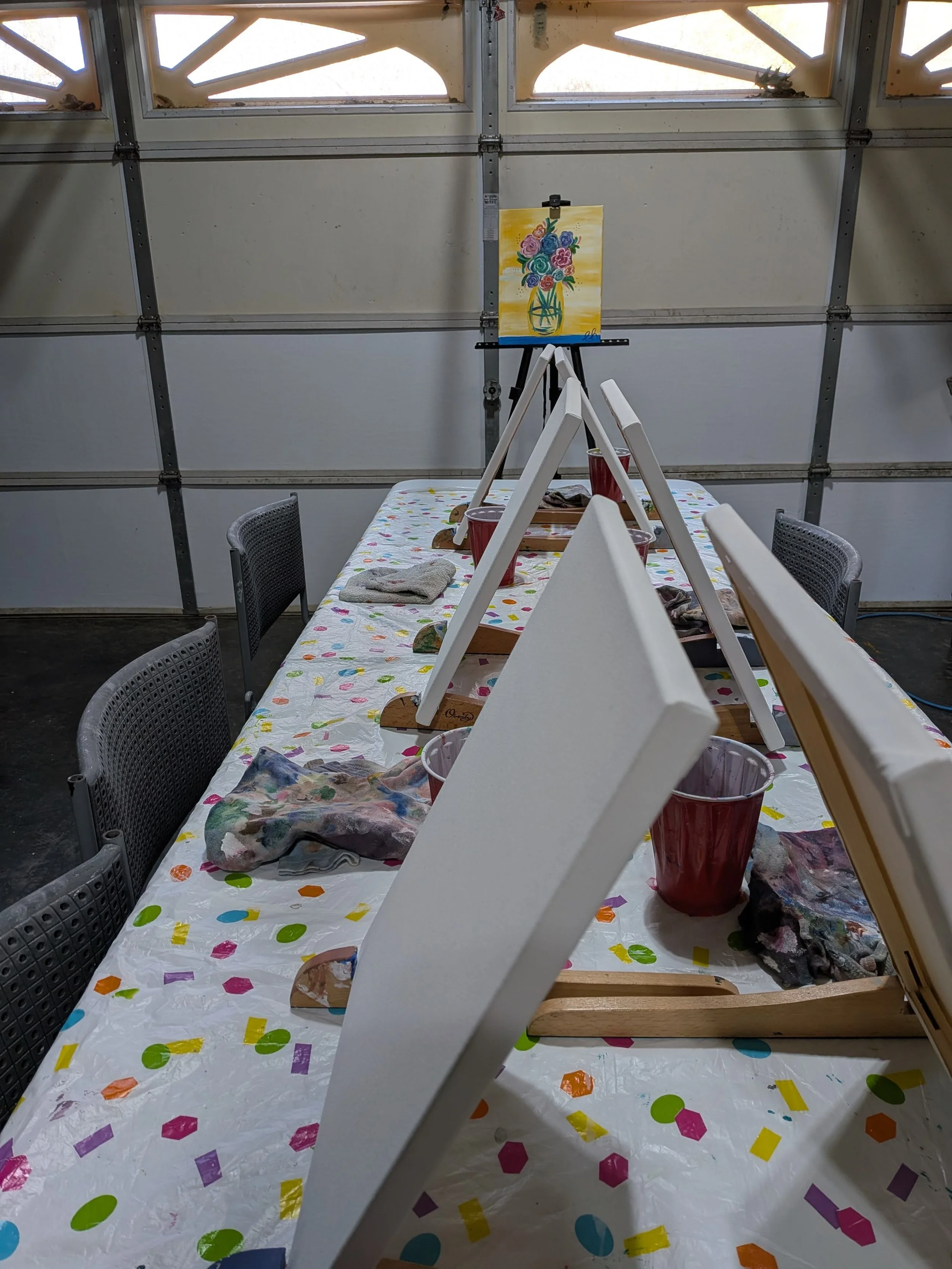 A long tabletop covered with colorful confetti-like paper, four small wooden easels, and various painting supplies, including buckets of red paint and towels. An easel at the end displays a painting of a bouquet of flowers in a yellow vase.
