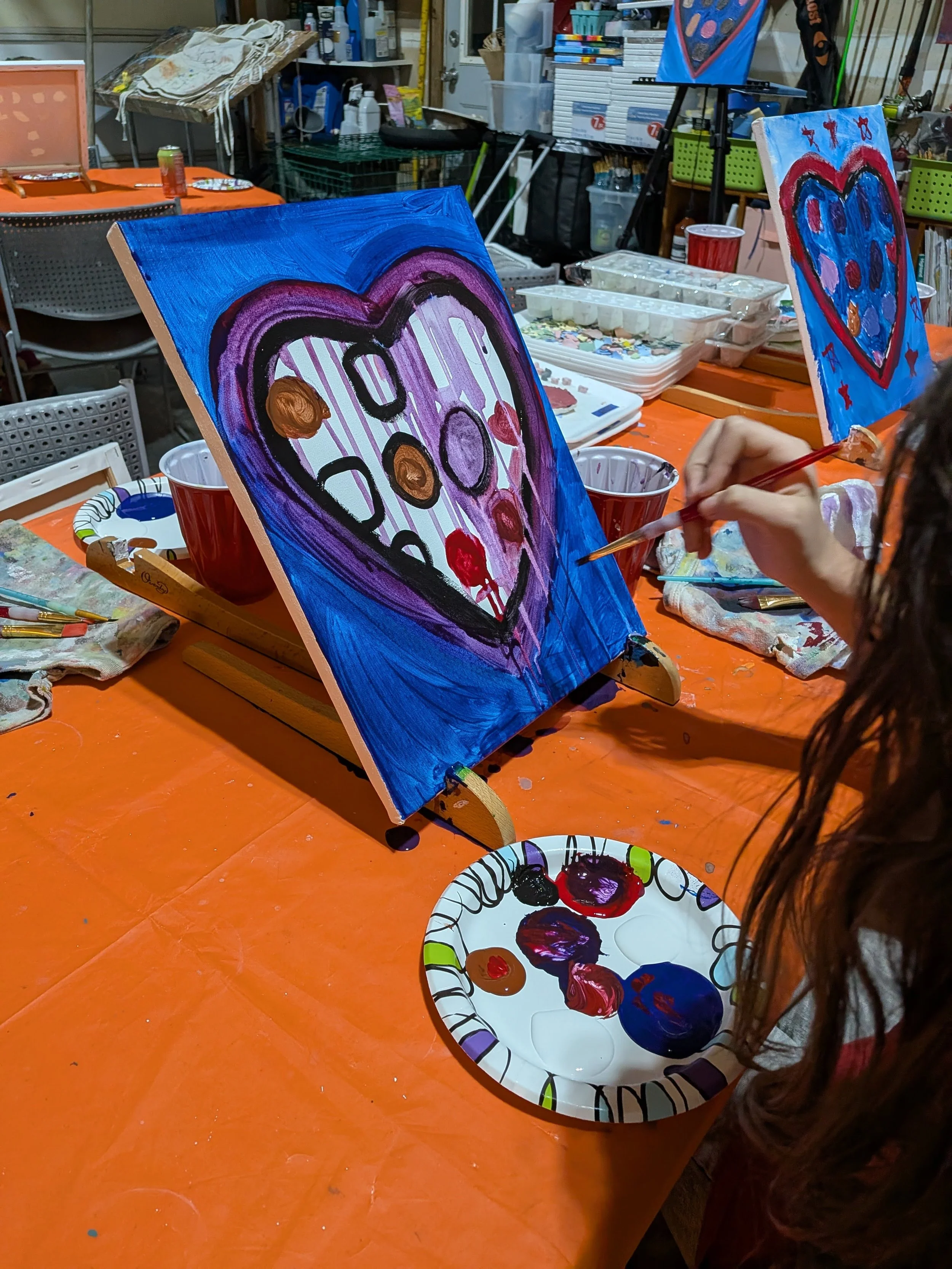 A person painting a colorful heart with a face on a canvas, sitting at a table covered with an orange tablecloth, surrounded by painting supplies and other artworks in progress.