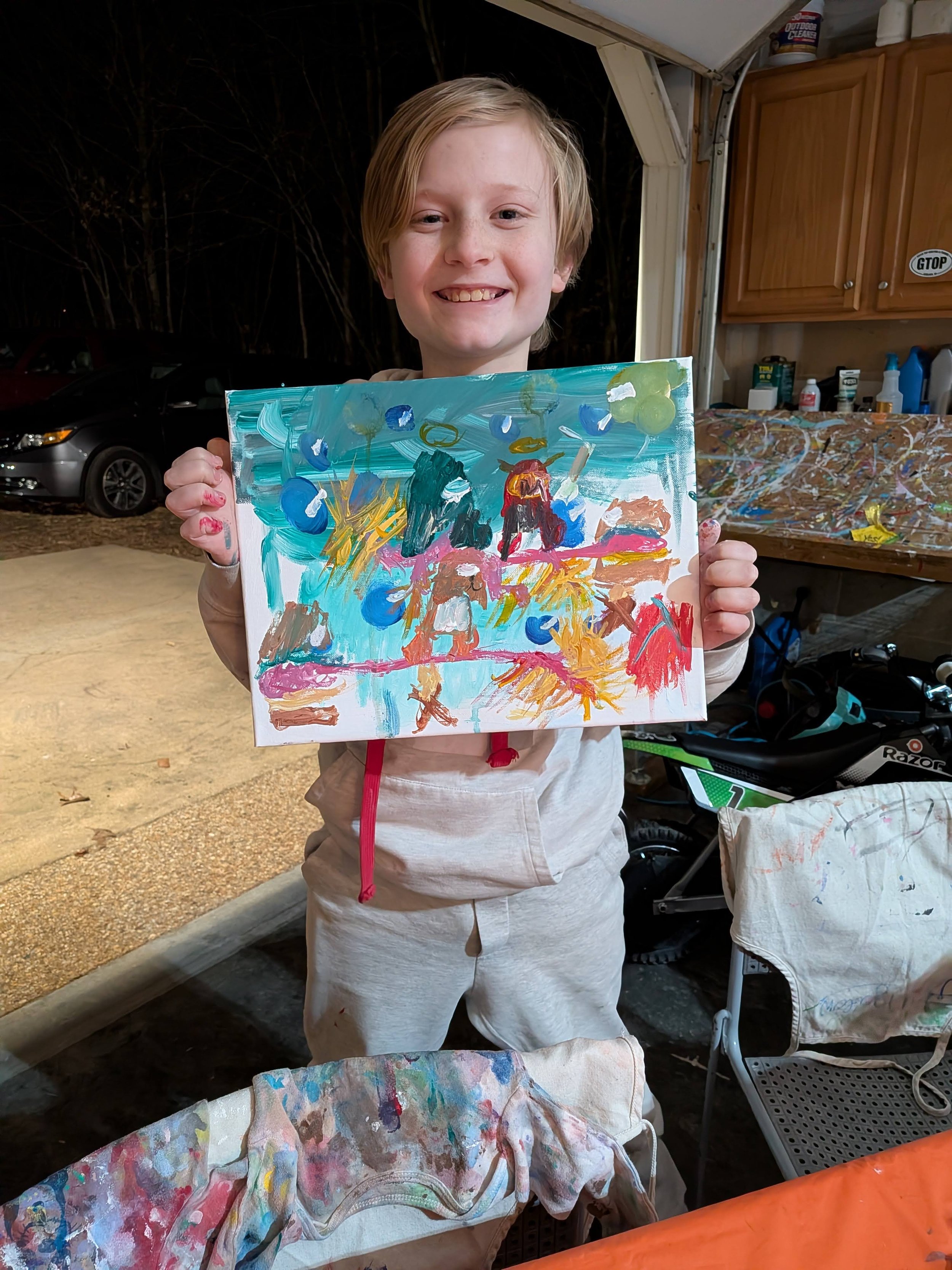 A young child with blonde hair smiling and holding a colorful abstract painting in an indoor or garage space.
