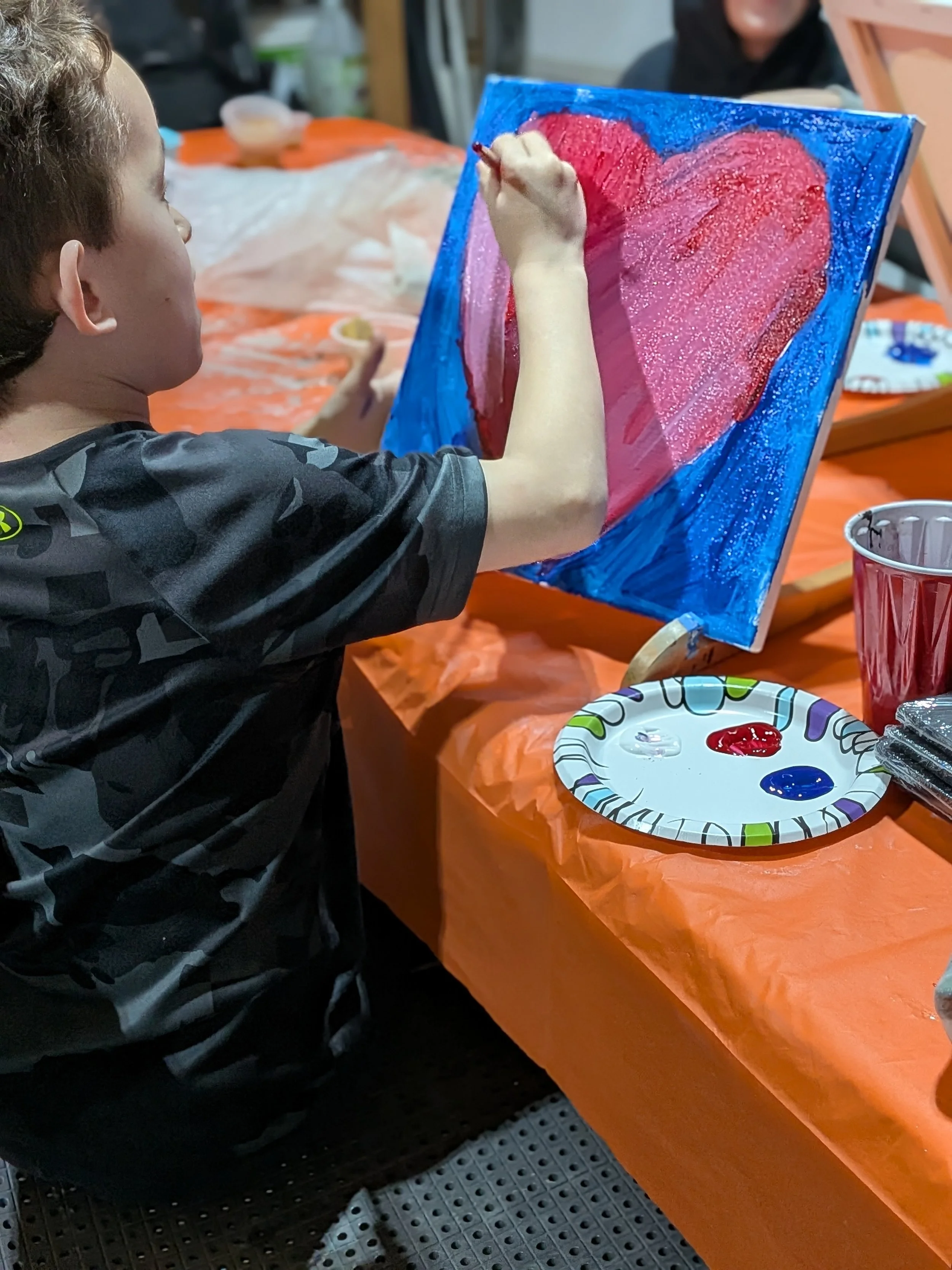 Child painting a large red heart on a blue canvas with pink highlights, sitting at a table covered with an orange tablecloth, with art supplies nearby.