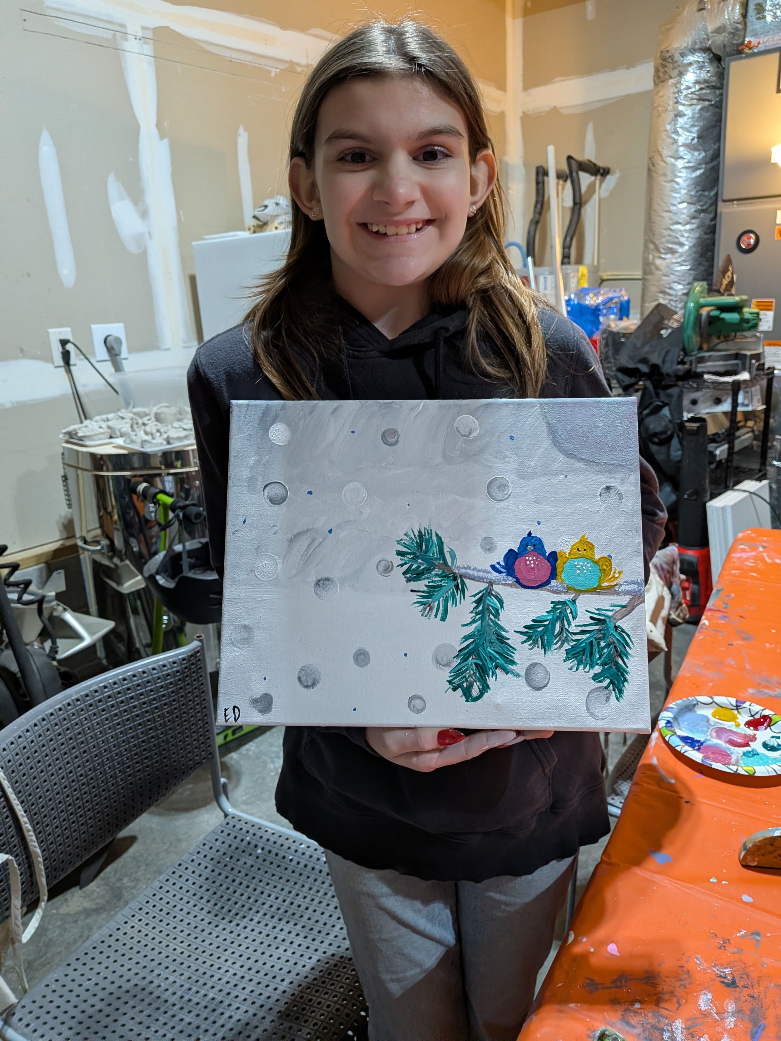 A girl holding a painted canvas depicting colorful birds sitting on a pine branch with a snowy background in a garage or workshop.