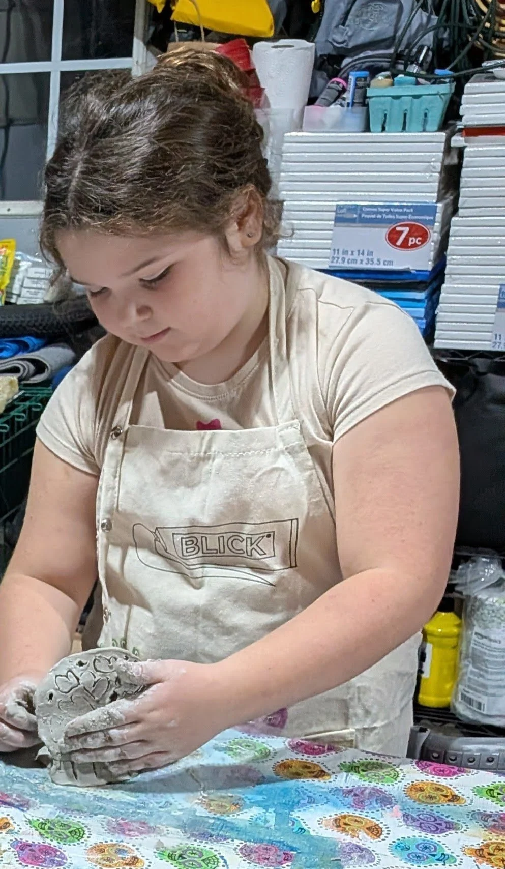 A young girl with brown hair tied back, wearing a beige T-shirt and a cream apron with the word 'BLICK' on it, is working with clay at a table covered with a colorful tablecloth.