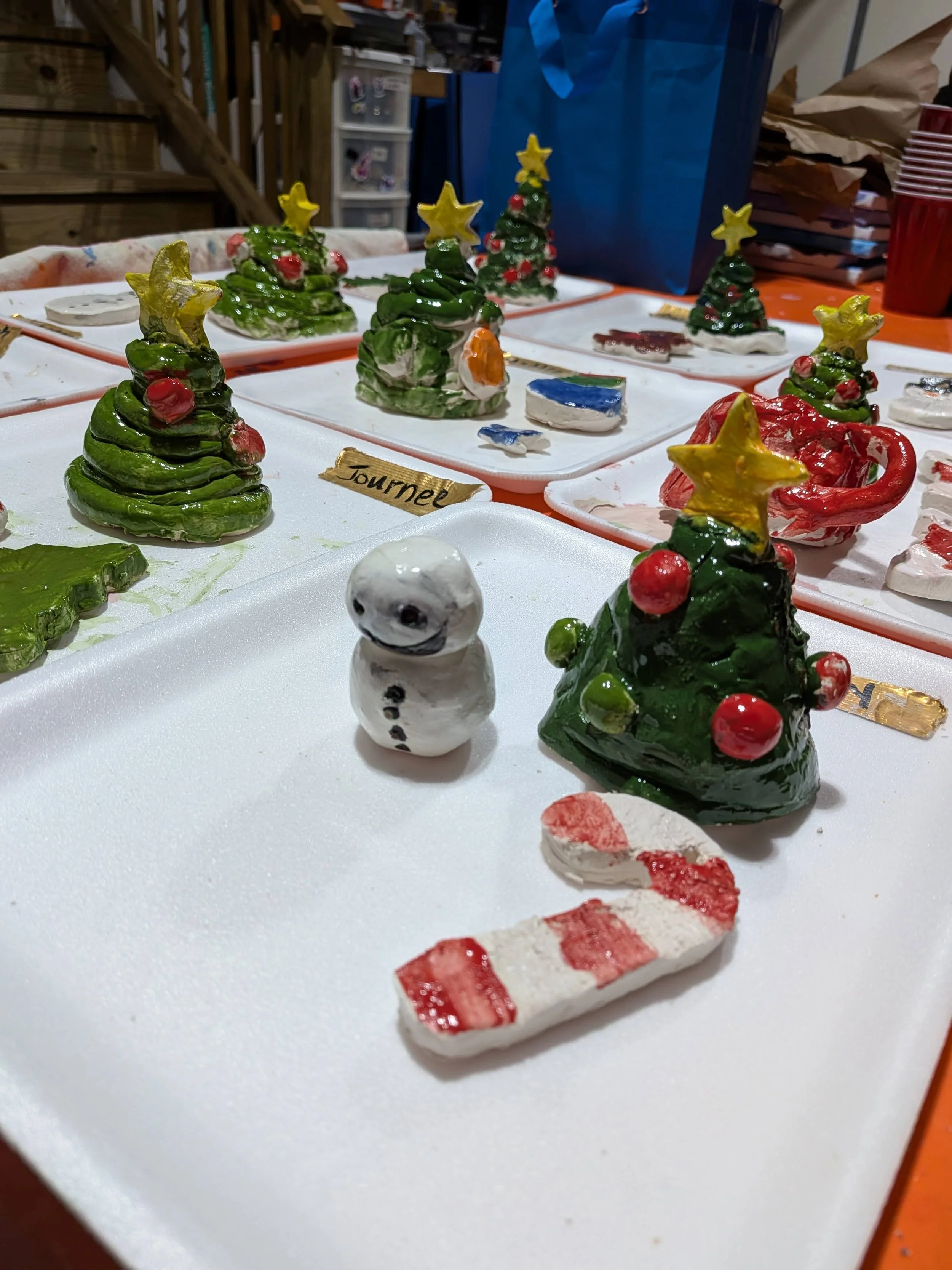 Display of handmade ceramic Christmas decorations, including Christmas trees, a snowman, and a candy cane, arranged on white trays.
