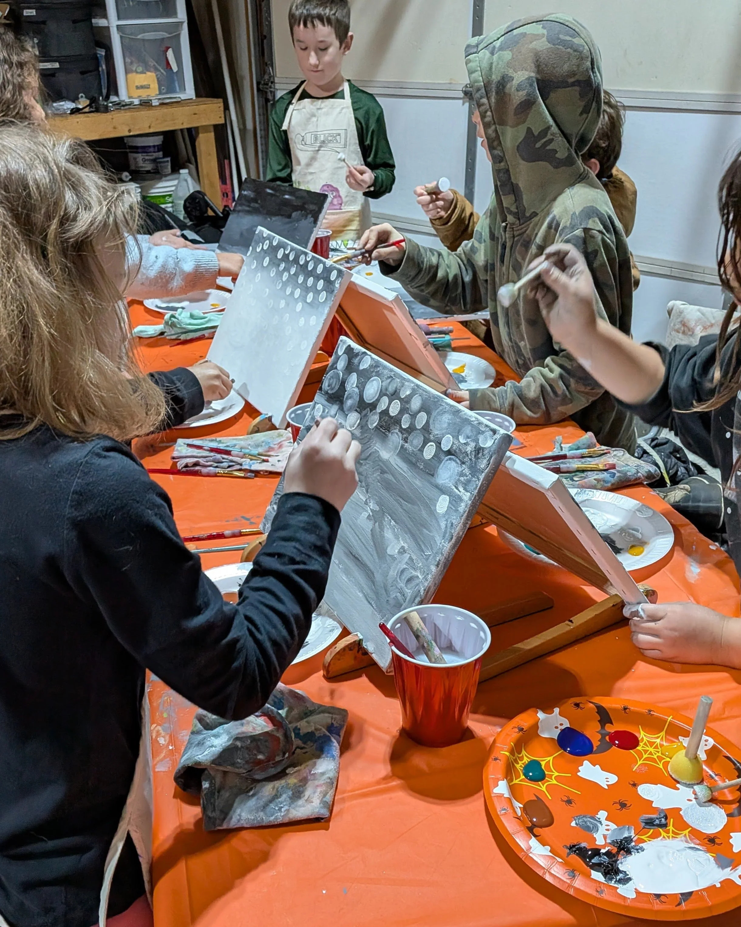 Children painting Halloween-themed canvases at a table with Halloween decorations and painting supplies.