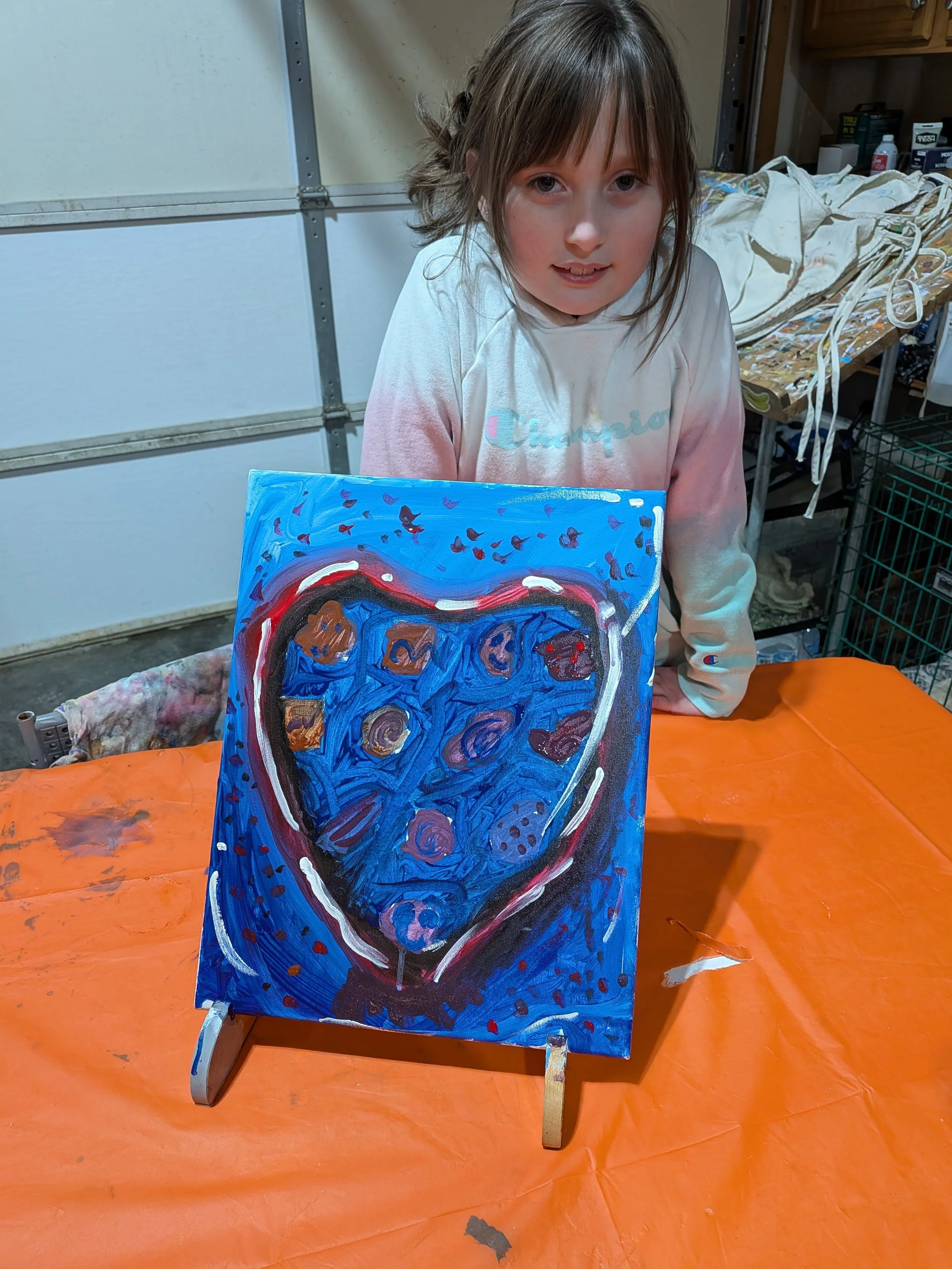 A young girl with long brown hair leans over a table showing a painted canvas of a blue heart with various patterns inside it. The girl is wearing a white and pink sweatshirt with the word 'Champion' on it, and the background shows a garage setting w