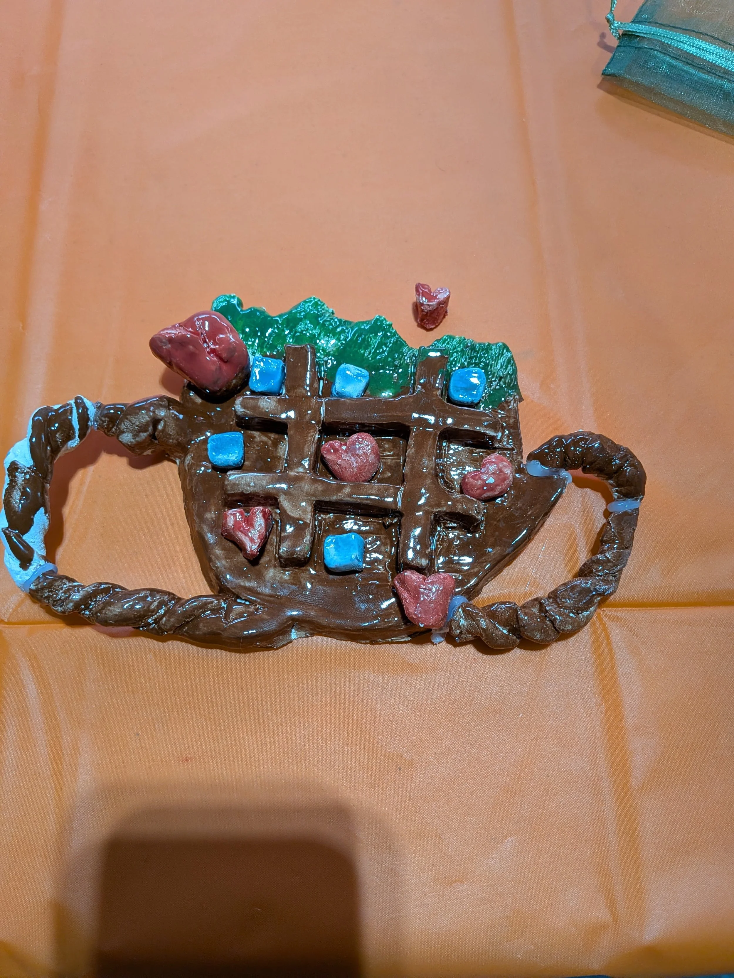 A teapot-shaped craft made from brown, green, and white materials, decorated with small red hearts and blue squares, and placed on an orange surface.