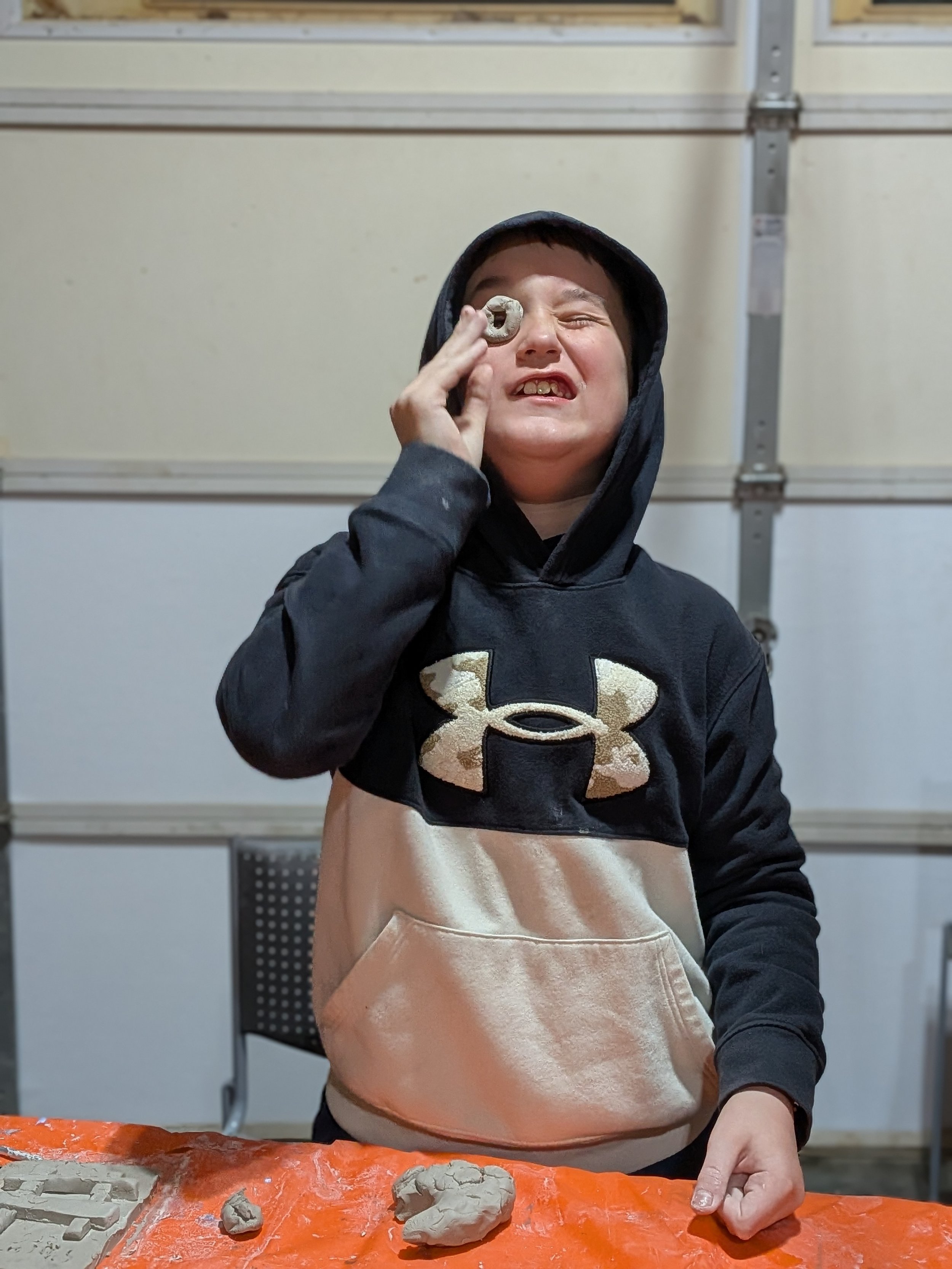 Young boy in a black hoodie with beige and black front pocket, holding a small clay object to his eye, with clay and molds on an orange table in front of him, in a garage or workshop setting.