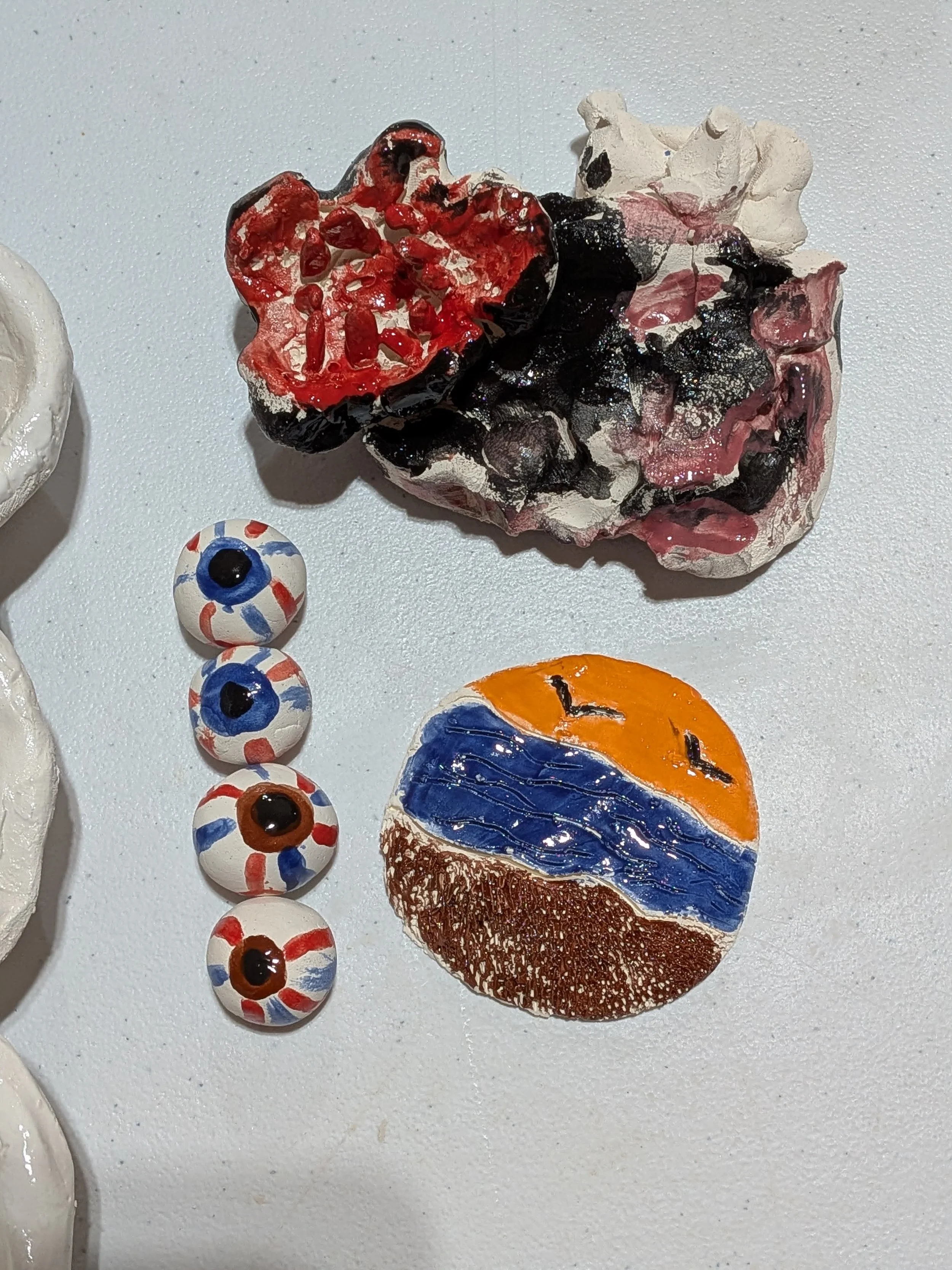 Colorful ceramic or glass art pieces with abstract designs, including an orange and blue circular piece with black 'W' shapes, a series of red, white, blue, and brown eyeball-like spheres, and a textured piece resembling a flower with red, black, and