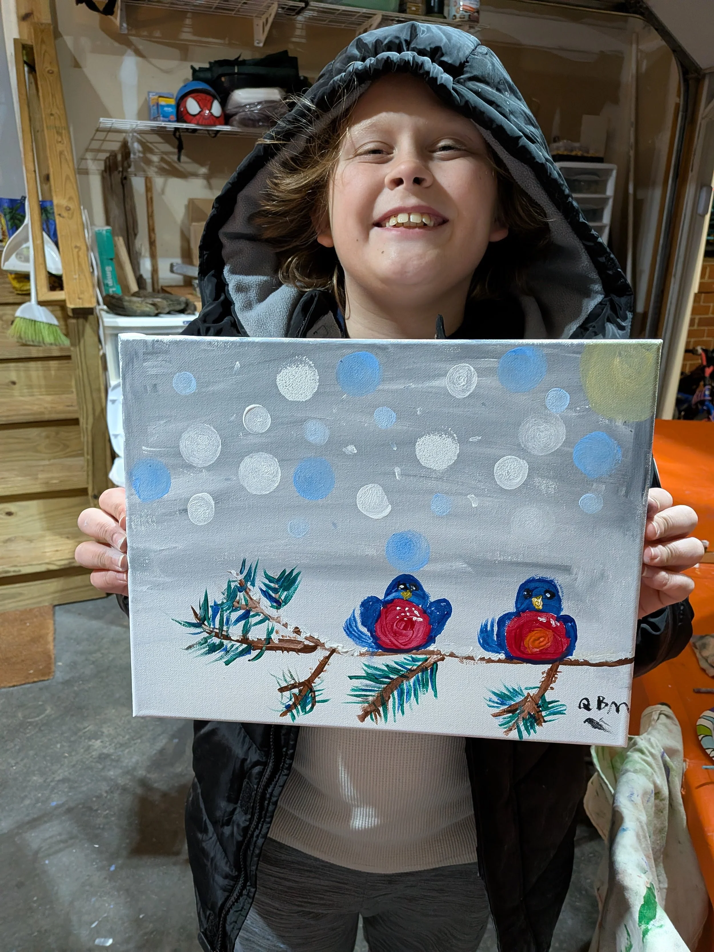 A young girl smiling and holding a painting of three blue birds perched on pine branches, with a background of blue and white circles, in a garage or workshop with shelves and tools.