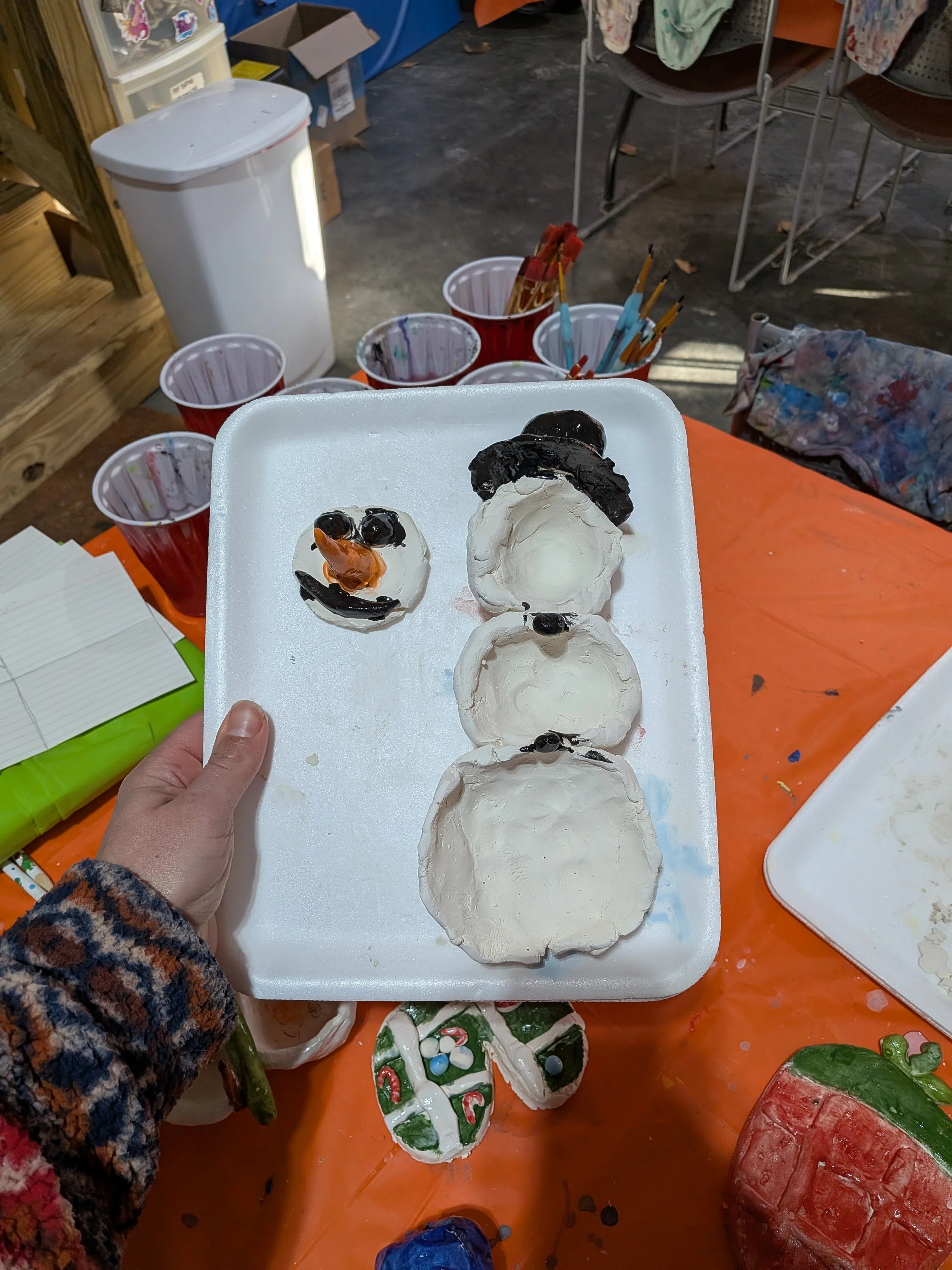 A person holding a foam tray with four decorated snowman cookies, each made from white icing with black and orange details, on a table covered with orange tablecloth. In the background, there are red cups with paintbrushes and art supplies, and a few
