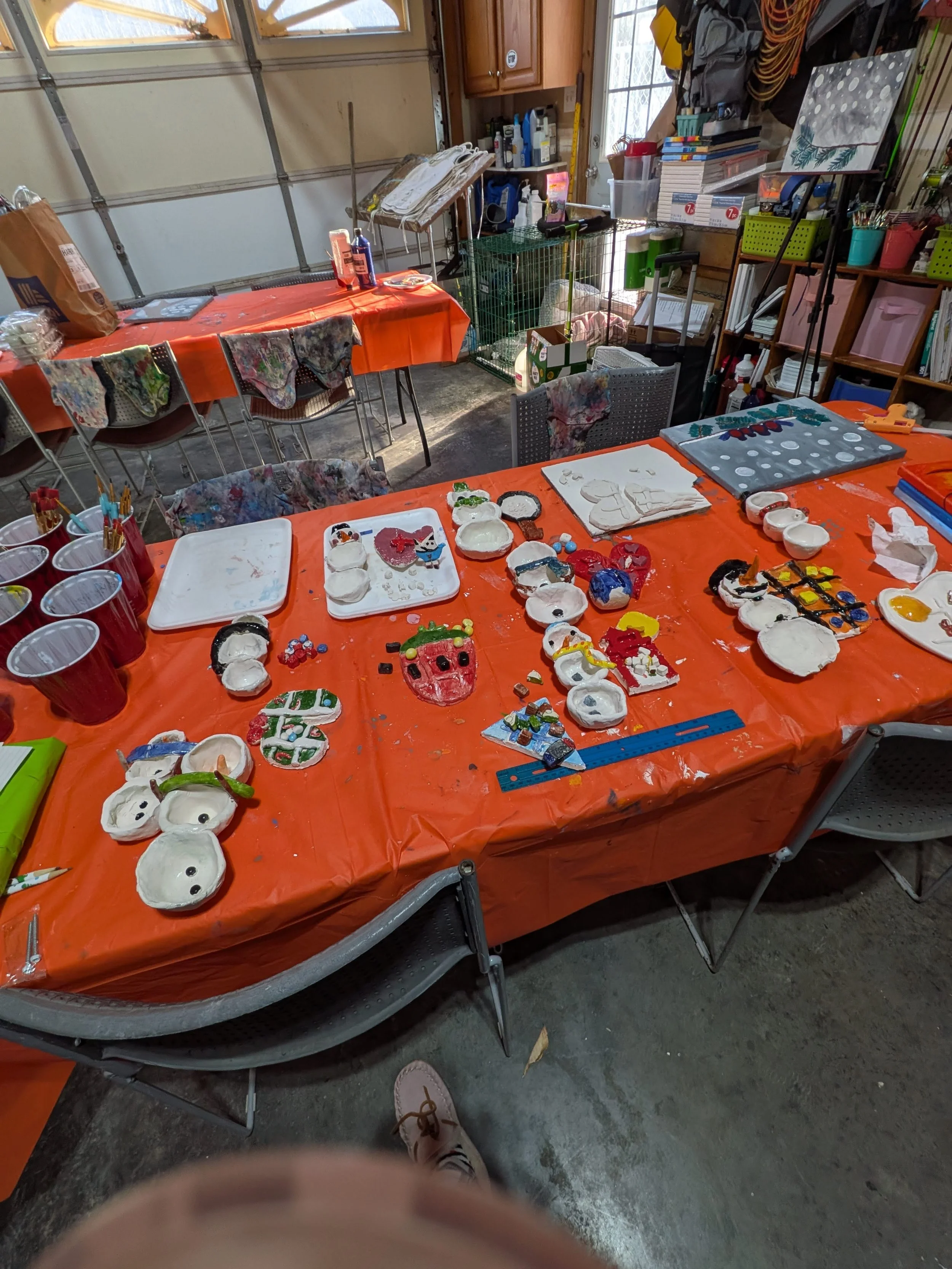 A garage workshop with an orange tablecloth covered table displaying painted ceramic ornaments and craft supplies.