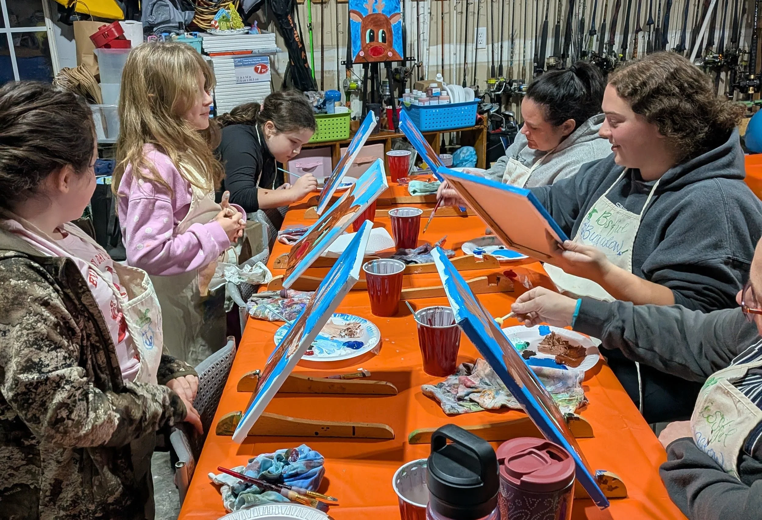 Group of people sitting at a long table painting canvases in a garage or workshop, with art supplies and tools in the background.