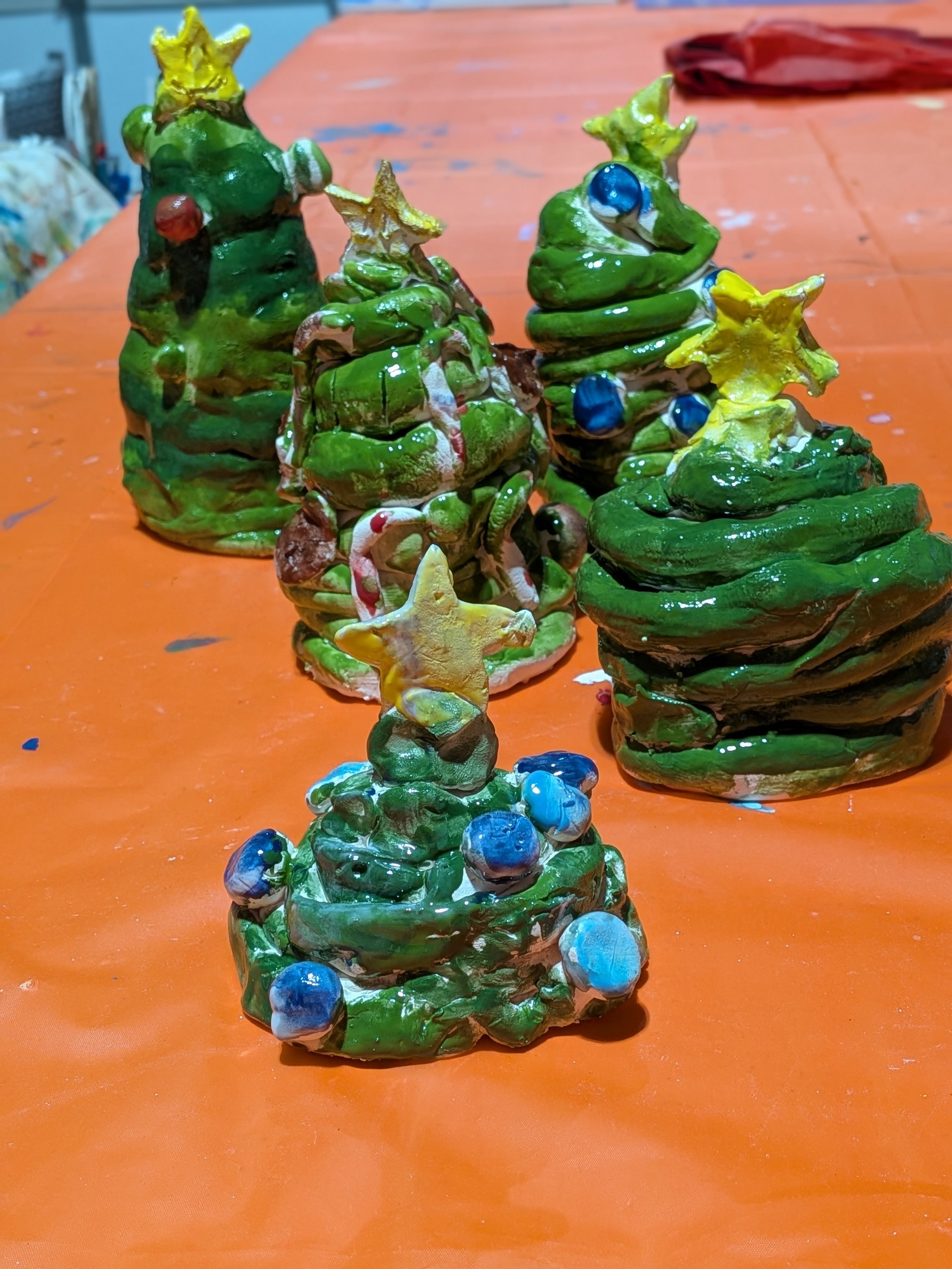 Handmade ceramic Christmas tree decorations painted green with colorful accents such as yellow stars, blue ornaments, and red berries, displayed on an orange surface.
