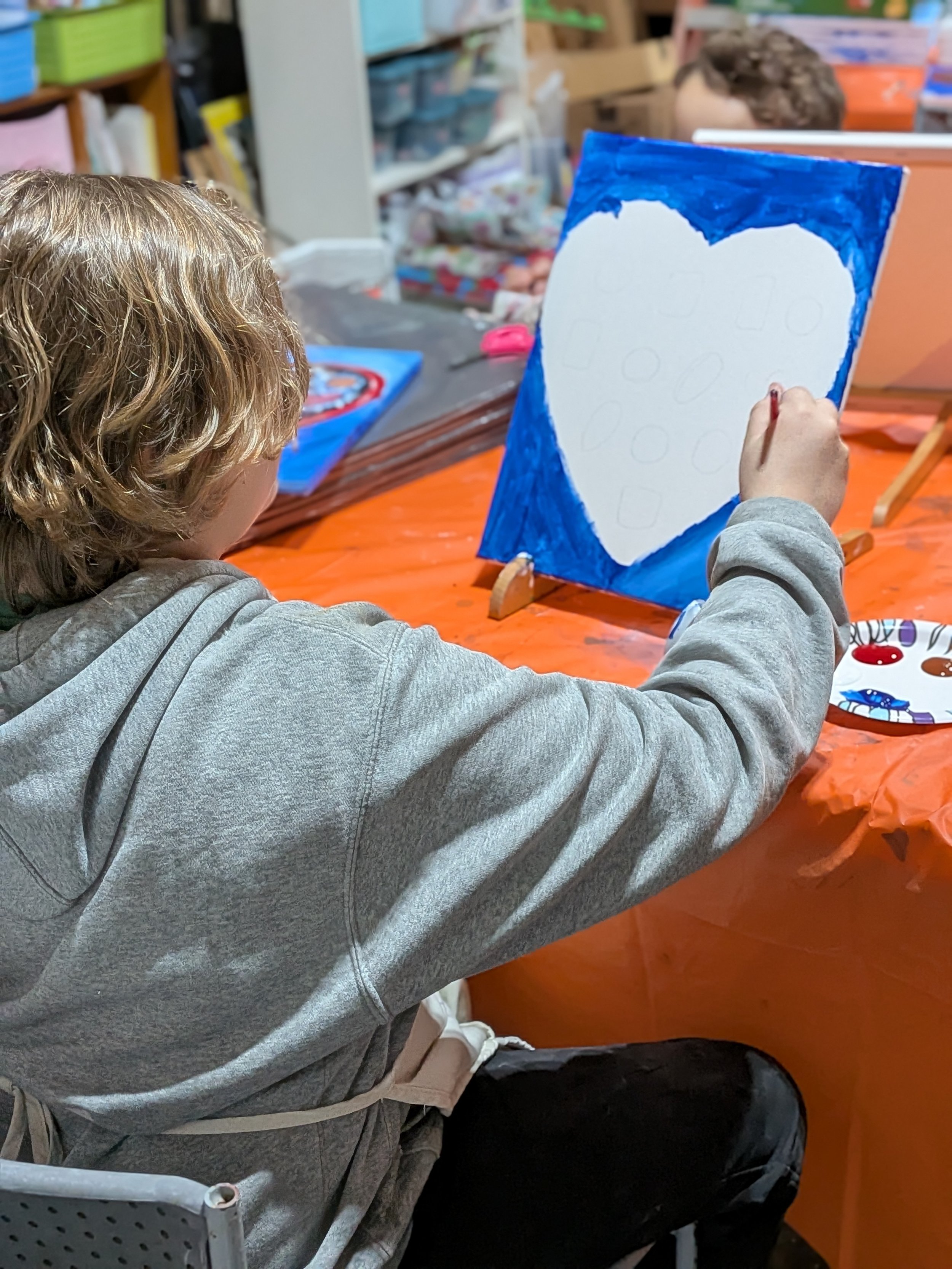 Child painting a large picture of a heart with a blue background at a craft table.