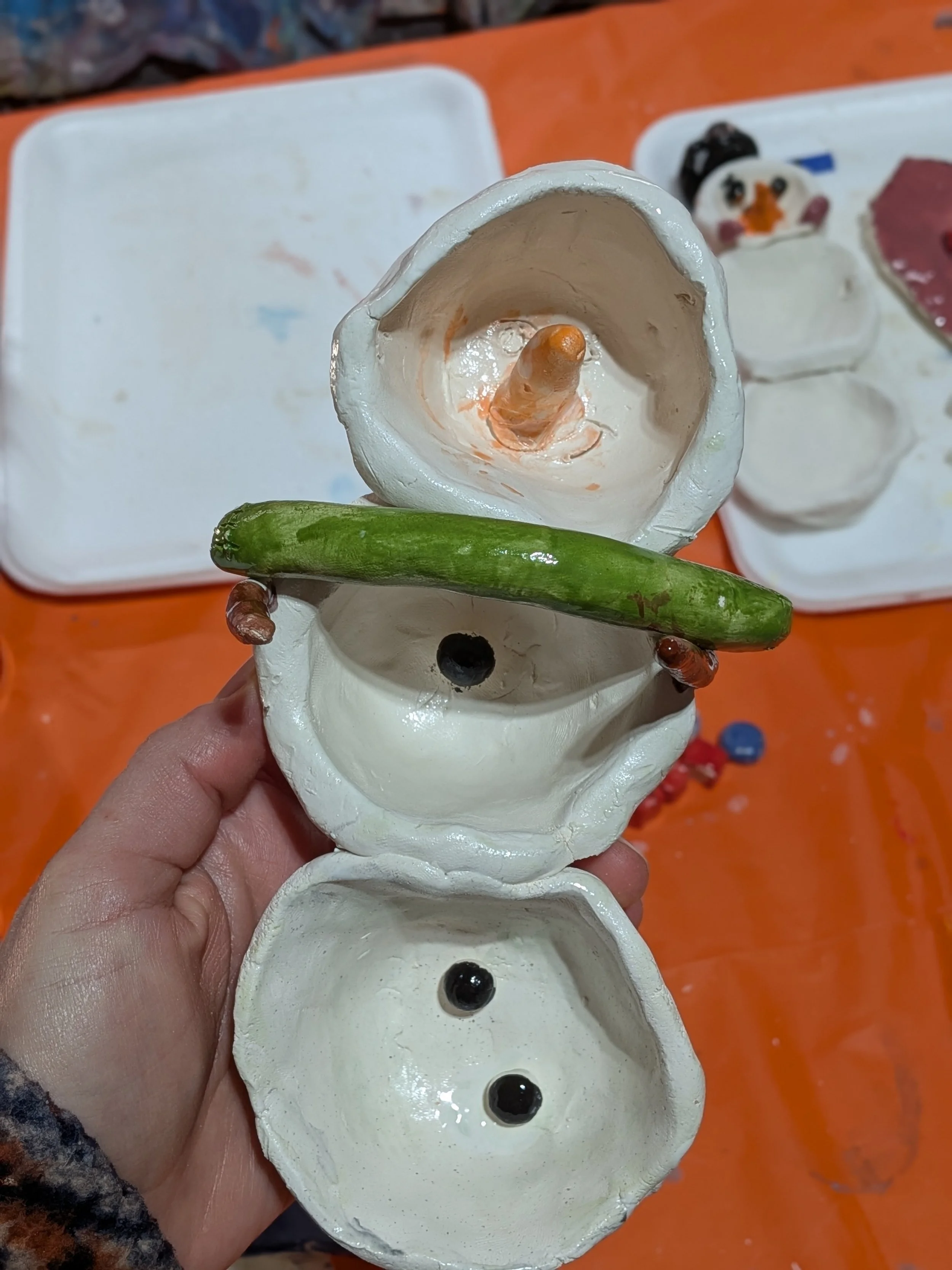 Ceramic snowman figurine with three open sections, holding a cucumber, with painted facial features. The background shows trays with additional ceramic figures on an orange surface.