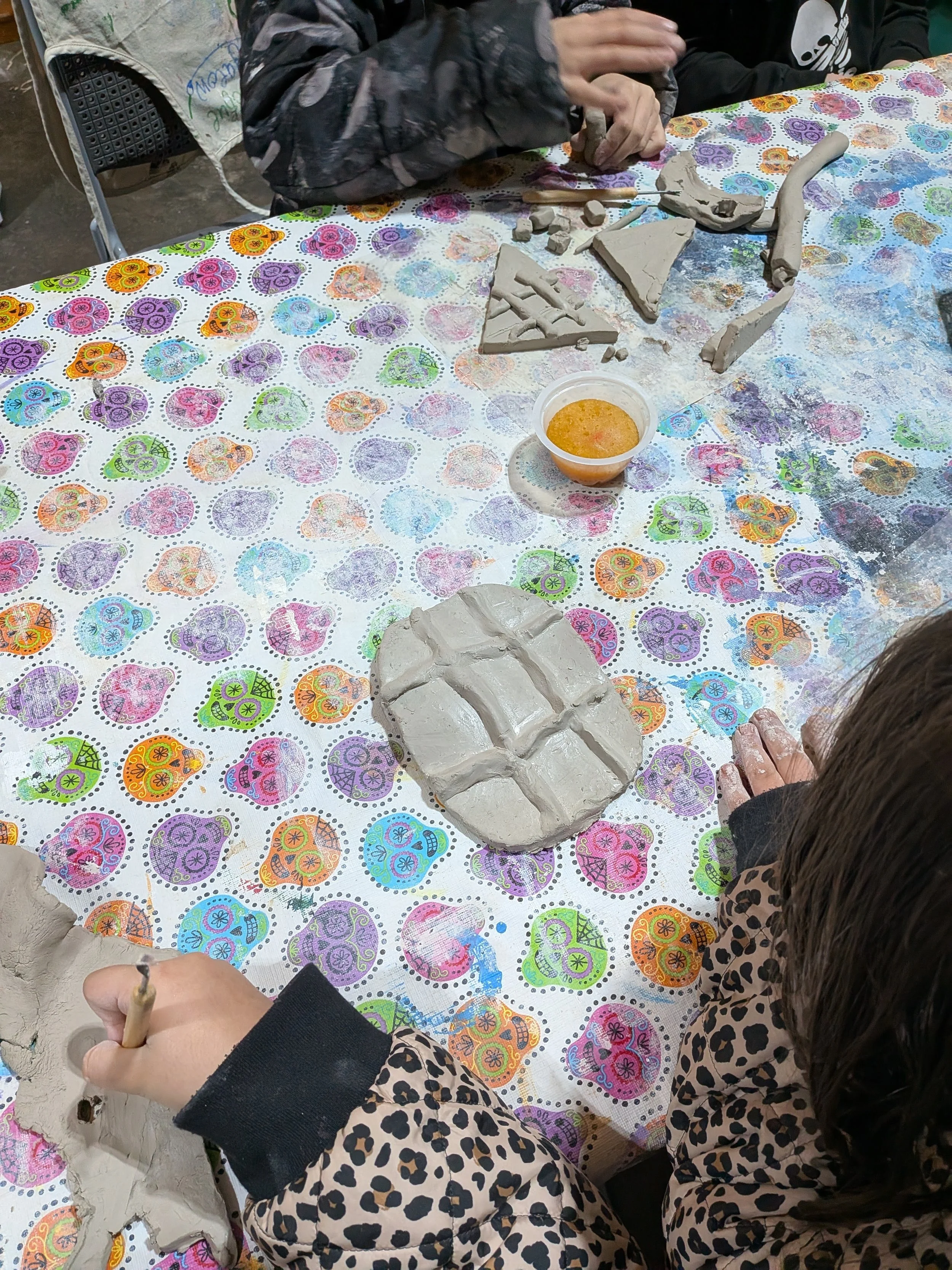 Individuals making clay sculptures at a table with a colorful, patterned tablecloth. There are clay pieces, tools, and a container of water or slip on the table.
