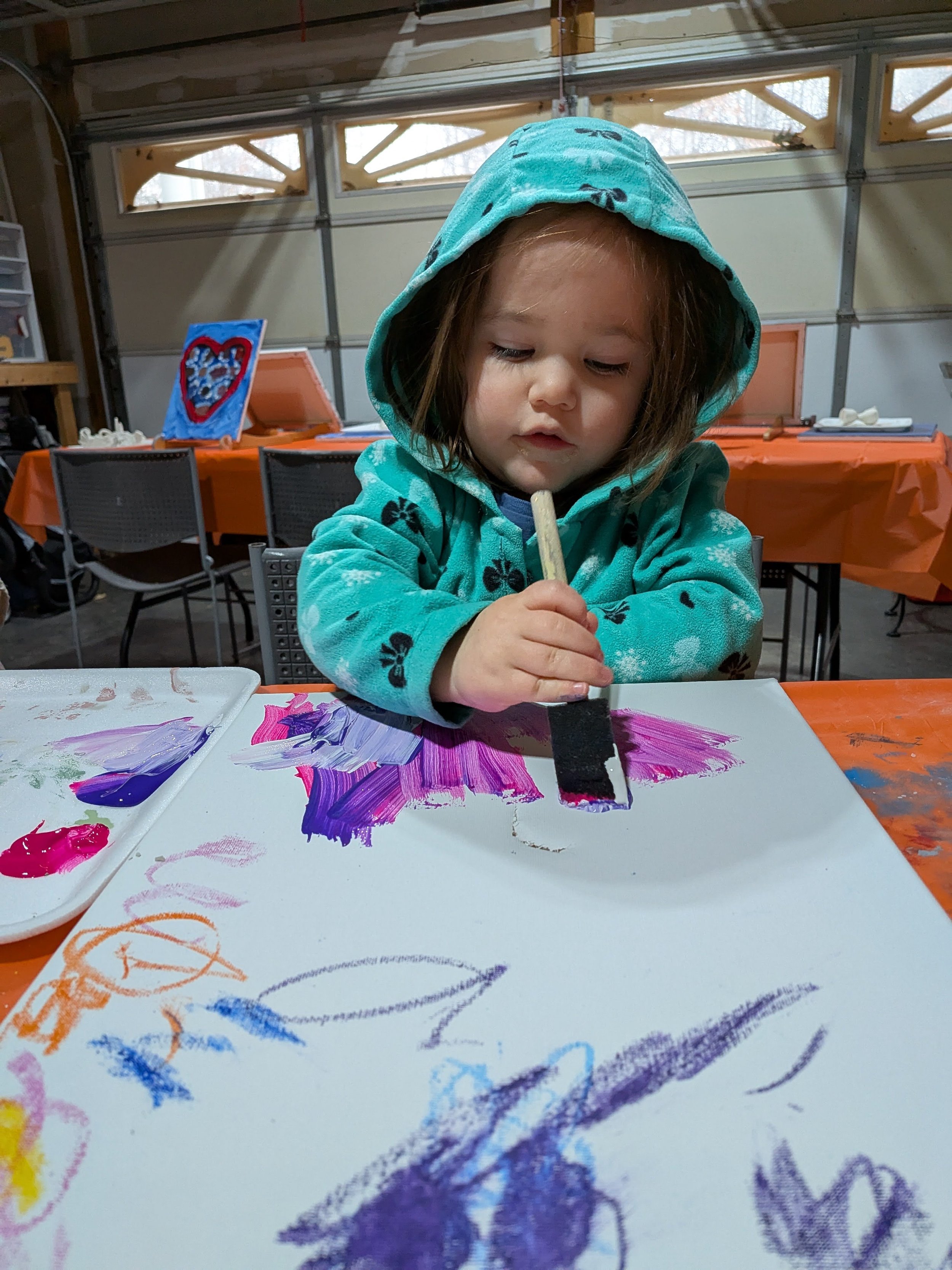 A young girl with a blue hoodie painting with a wide brush on a large white sheet of paper at a table in a garage or craft room.