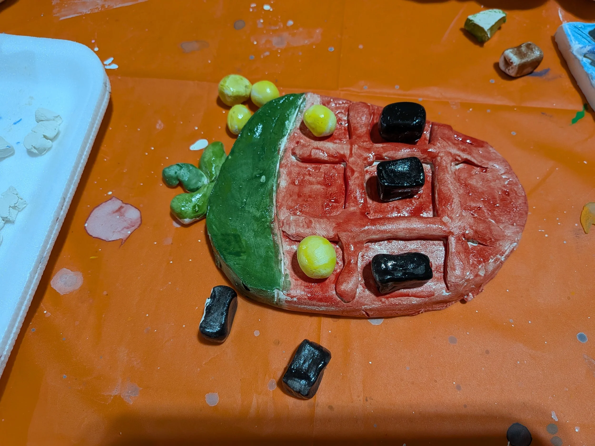 A colorful, watermelon-shaped craft made of clay with black, yellow, and green elements, on an orange table, with scattered small clay pieces around.