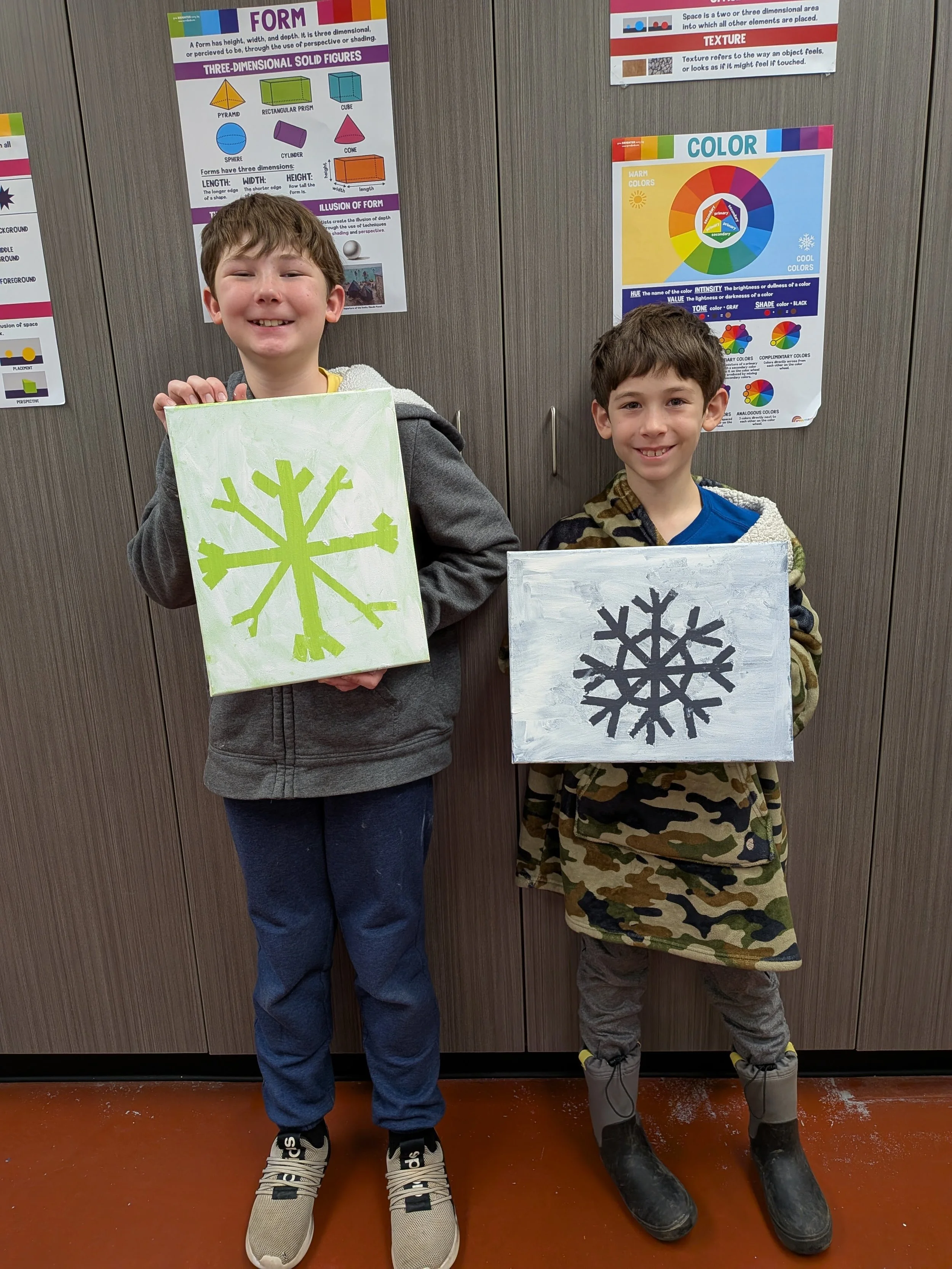 Two boys standing indoors against a wooden wall holding painted canvas art; one with a green snowflake on a light background, the other with a black snowflake on a gray background. They are smiling.