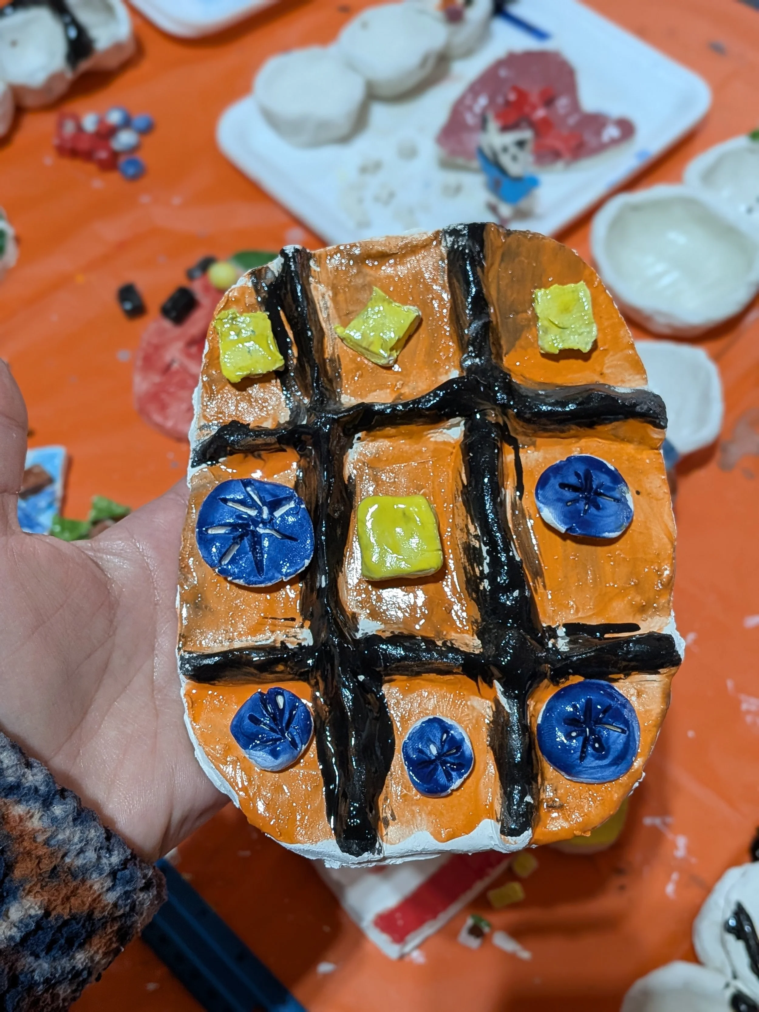 A decorative cake resembling a tic-tac-toe board, with orange icing, black lines, and blue and green fondant pieces on it.