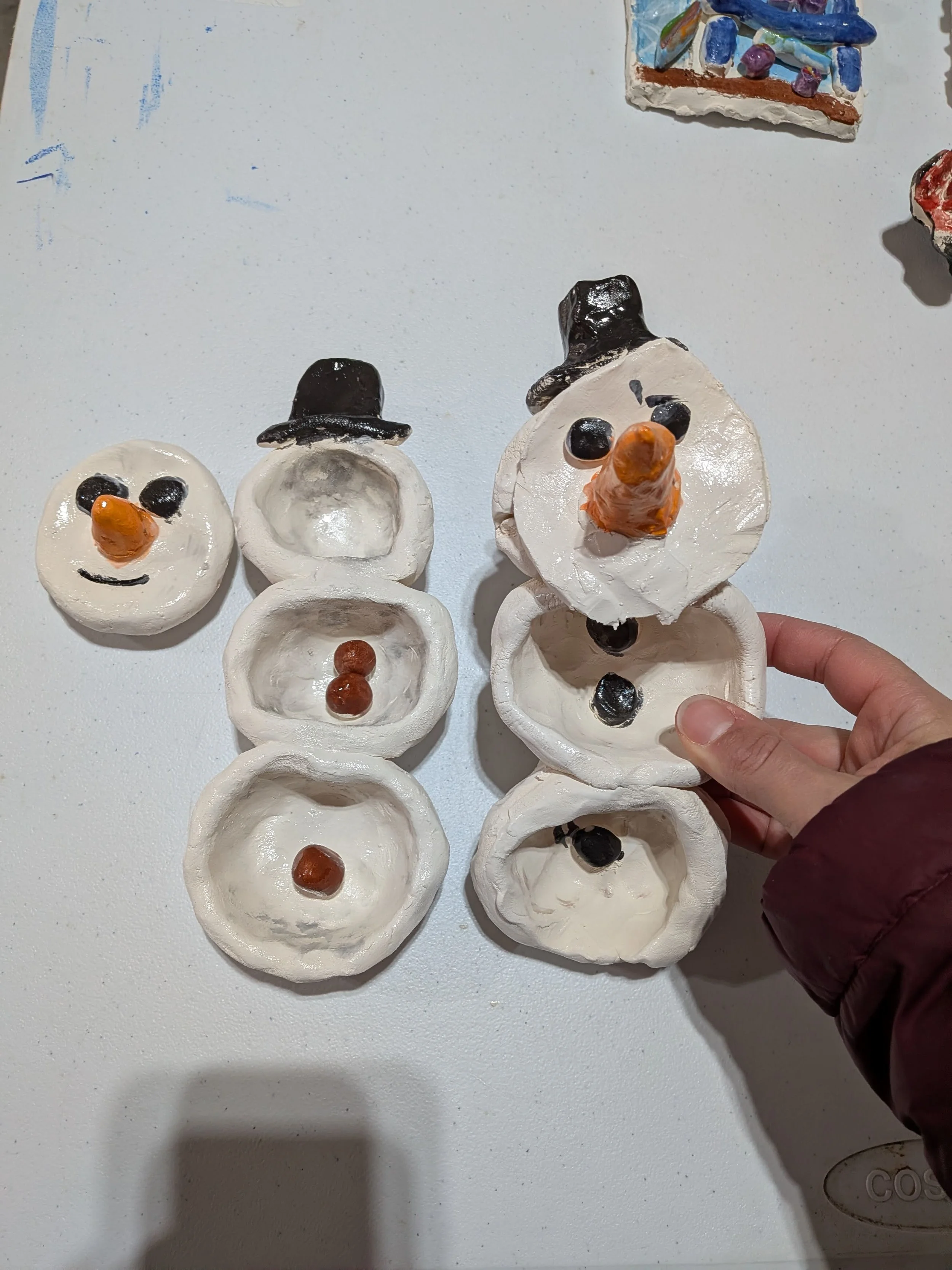 Handmade snowman craft project made from painted egg cartons, with carrot-shaped noses, black eyes, and black hats, arranged on a white surface.