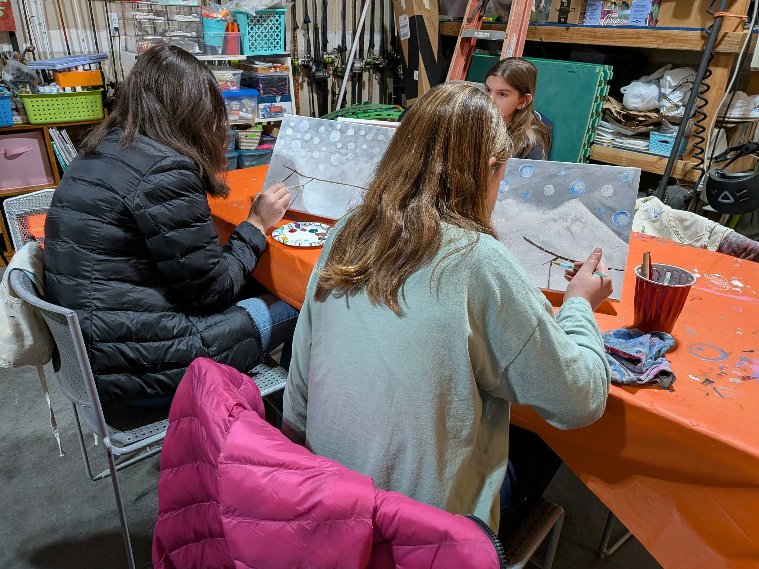 Three young women painting canvases at a table covered with an orange tablecloth in an art studio. The canvases depict a snow scene with a mountain and snowball-like circles in the sky. The workspace is filled with art supplies and storage shelves.