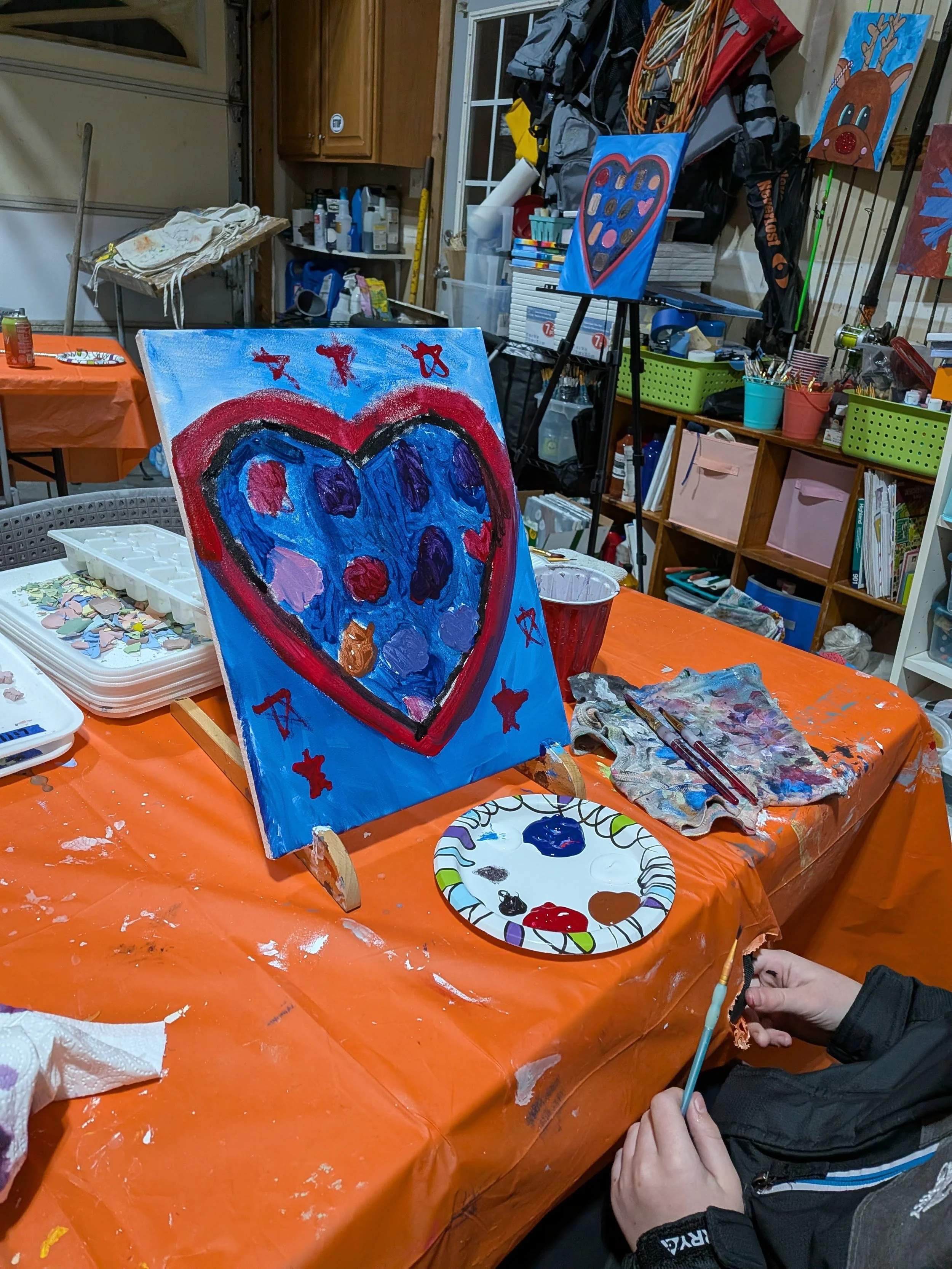 A young person painting a colorful heart on a canvas in an art studio. The canvas features a large blue heart with purple and red spots, surrounded by red stars and a red outline. The person holds a paintbrush and is working on the painting, which is